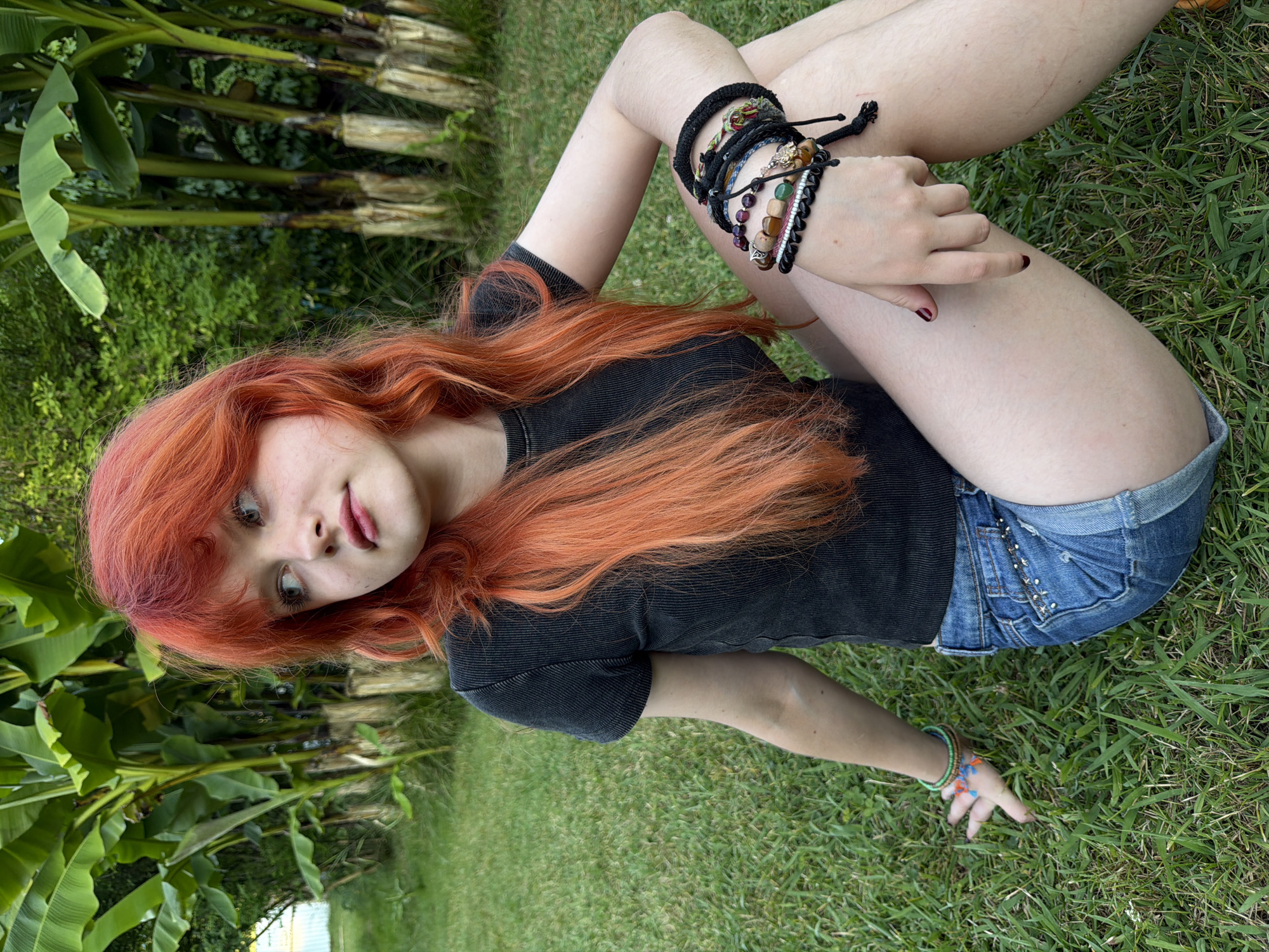 This media file features a young woman with vibrant red hair lying on a grassy lawn, likely in Sochi, Russia given the context. She is wearing a black t-shirt and denim shorts. Her pose is relaxed, with her legs bent and her arms resting on the grass. She has multiple bracelets on her left wrist, featuring a variety of beads and charms, and a lighter, colorful bracelet on her right wrist. The background shows lush green foliage, possibly banana trees, and tall, slender plants, suggesting a tropical or sub-tropical environment, consistent with Sochi's climate. The lighting indicates it is daytime, with natural sunlight illuminating the scene. The woman's expression is somewhat playful, with a slight smile and direct gaze towards the camera, conveying a sense of ease and enjoyment of the outdoor setting. There is no visible text in the image.