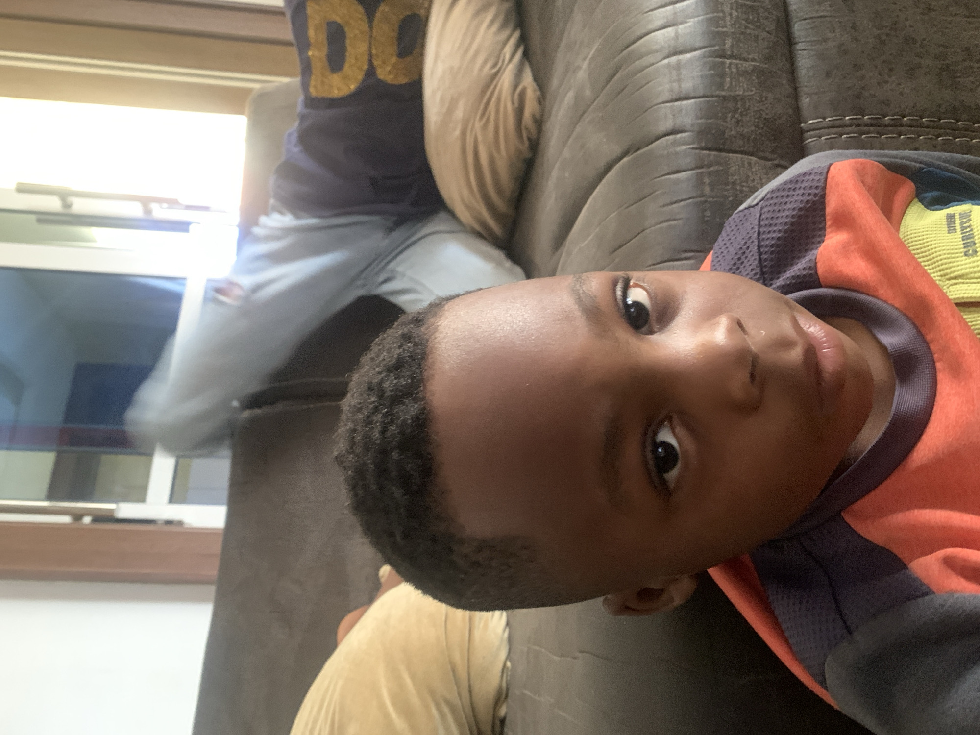 A young boy is lying on a brown leather couch, looking towards the camera. He is wearing a bright orange and yellow shirt with dark gray sleeves. Behind him, another person is partially visible wearing a dark blue shirt with gold lettering and light blue jeans. This person is sitting in a beige armchair or part of the couch. The background shows a window with partially visible blinds and light coming through. There is no information in the image to confirm this image was taken in Jos, Nigeria.