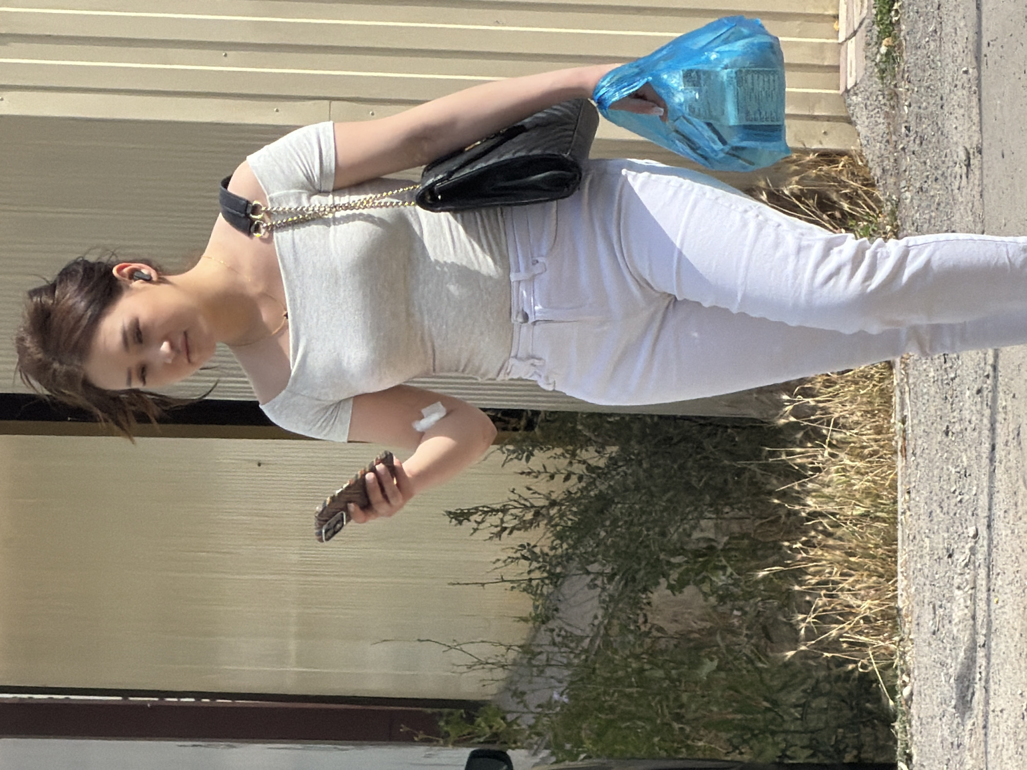 A young woman with dark hair pulled back is walking outdoors, looking at her smartphone, which she holds in her right hand. She is wearing a light gray off-the-shoulder top, white jeans, and white sneakers. A black quilted purse with a gold chain strap is slung over her left shoulder. In her right hand, she also carries a bright blue plastic bag containing items. She has an earbud in her left ear and a white bandage on her right upper arm. The background consists of a textured wall and dry, sparse vegetation.