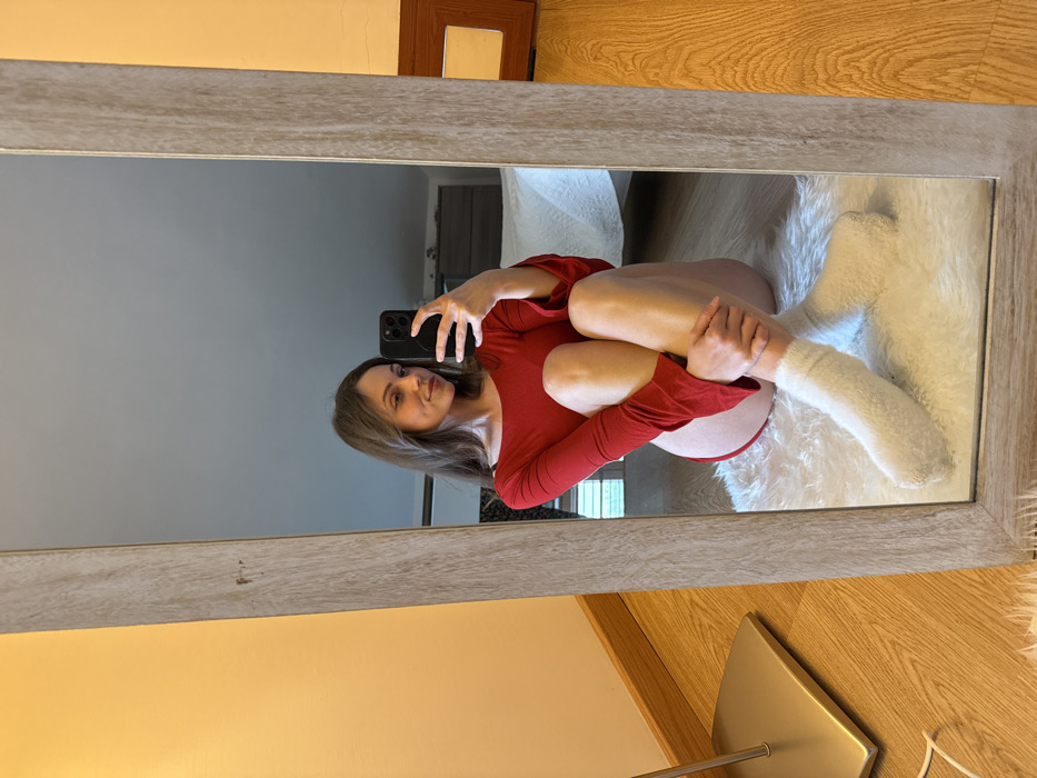 An adult female is depicted in an indoor mirror selfie. She is seated on a white, shaggy rug, facing her reflection in a full-length mirror featuring a light wood-grain frame. She wears a long-sleeved red top and white, fuzzy socks. Her legs are drawn towards her body, and her left hand rests on her right knee. In her right hand, she holds a dark-colored smartphone, equipped with a circular pop-socket, pointed towards the mirror to capture the reflection. The background visible in the mirror shows a room with a bed featuring white bedding, a dark dresser, and a section of light-toned wooden flooring. To the immediate left of the mirror, a metallic lamp base is visible on a matching wooden floor. A wall section to the left of the mirror is painted a muted yellow-orange. The precise location within Cava de' Tirreni, Italy, is not identifiable from the visual information provided.