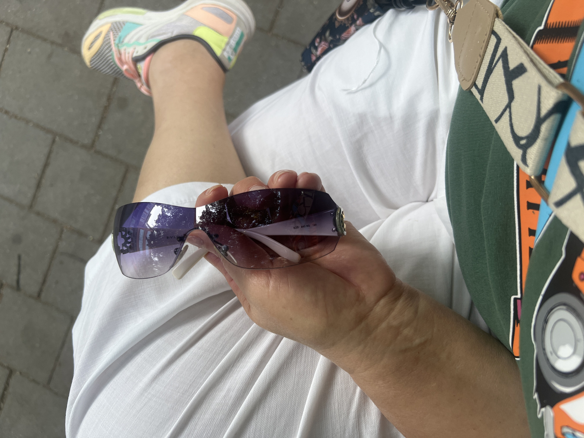 The image shows a close-up of a person's hand holding a pair of sunglasses. The sunglasses have purple lenses and a reflective surface that shows a building. The person's hand is fair-skinned with long fingernails. They are wearing a white garment and a green shirt with a strap from a bag visible at the bottom. In the background, there is a paved surface made of grey rectangular tiles. A person's leg and foot, wearing a colorful athletic shoe, are visible on the right side of the image. The overall impression is of a person sitting outdoors, likely in a public space given the paving and the presence of a bag. The image does not contain enough specific details to determine the exact location within the city of Bender, Moldova.