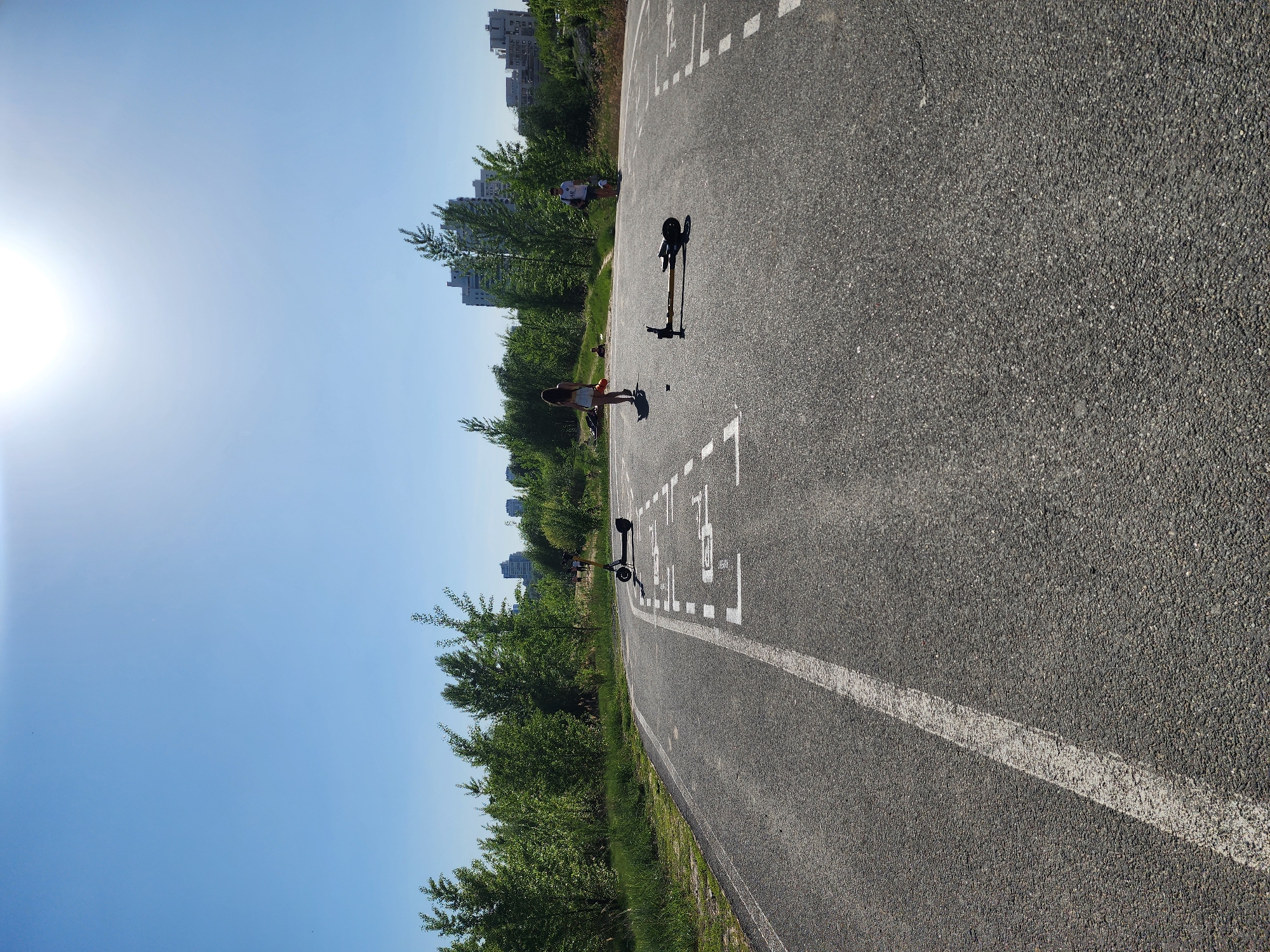 A horizontal outdoor shot of a paved road, likely in Kazan, Russia, shows a path with white markings and Chinese characters. The sky is clear and light blue. Buildings and trees flank the road. Several people are visible along the road's edge, including one woman in light-colored shorts and a tank top and a man in a white t-shirt and dark shorts. Three scooters are standing on the pavement. The sun is bright in the upper left corner.