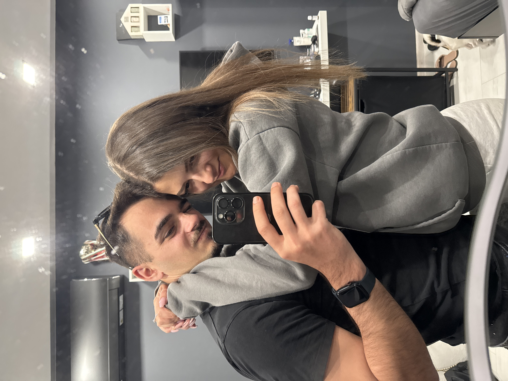 A smiling couple takes a mirror selfie in a modern indoor setting. The man, in the foreground, holds a black smartphone with his right hand to capture the image. He has short dark hair, a mustache, and a beard, and wears a black t-shirt. Sunglasses are perched on his head, and a dark smartwatch is visible on his left wrist.
Behind him, a woman embraces him, resting her head on his shoulder or chest. She has long brown hair and is smiling warmly. She wears a grey hooded sweatshirt, and her left hand, adorned with painted nails, is seen wrapped around the man's waist.
The background, reflected in the mirror, shows a minimalist interior. A dark grey wall is visible behind the couple, featuring a small white decorative miniature house mounted on it. To the right, a black clothes rack or piece of furniture can be seen, with a pair of light-colored shoes on the floor nearby. The overall ambiance suggests a comfortable and affectionate moment. No explicit text is visible in the image. The scene takes place in Sofia, Bulgaria.