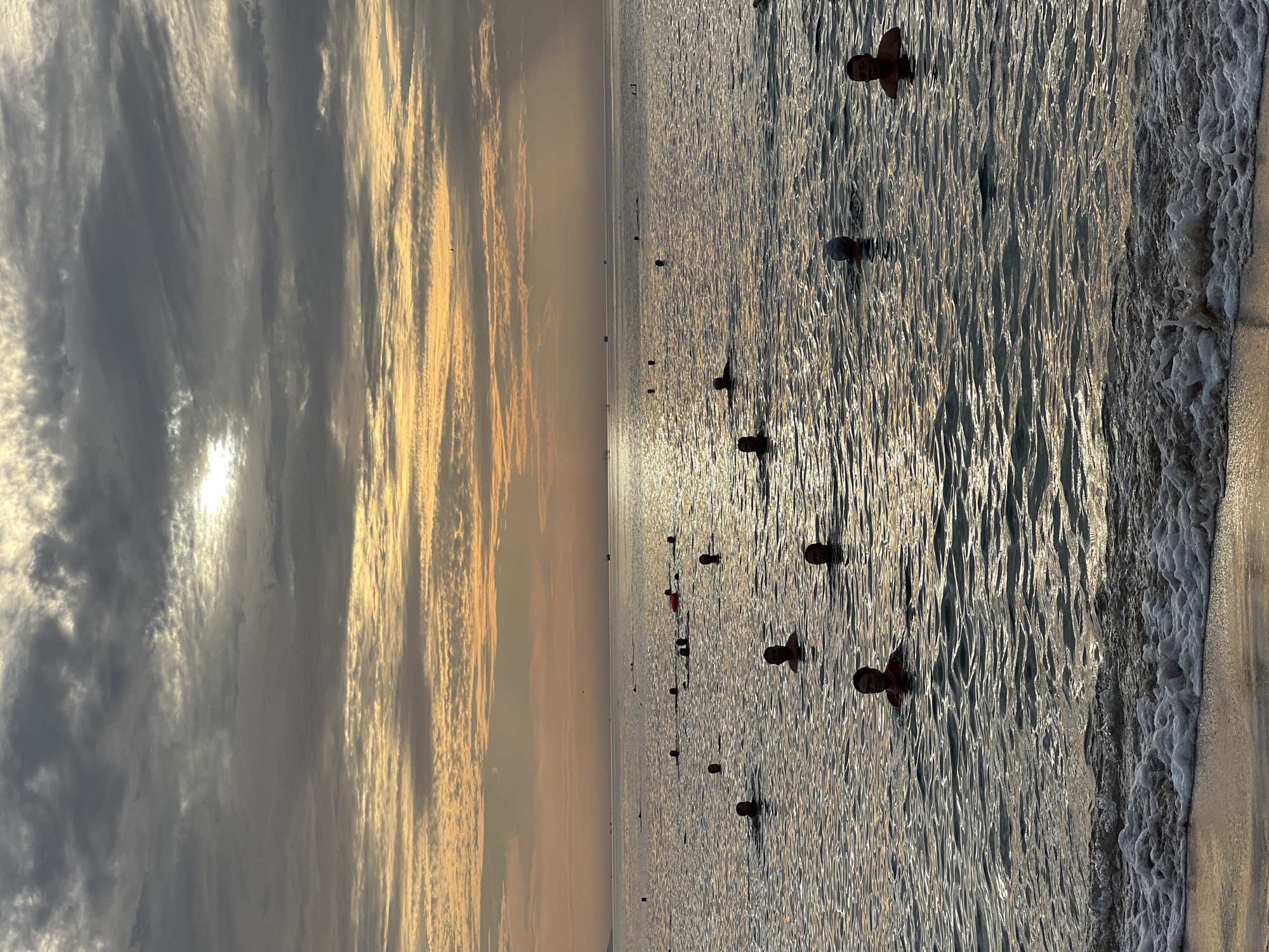 The image depicts a group of people swimming in the ocean at Nha Trang, Vietnam, during a cloudy sunset. The sun is visible through a break in the clouds, casting a warm glow on the water. The waves are gentle, with foam lapping at the sandy shore in the foreground. The people are scattered across the water, some with their heads just above the surface, while others are further out. The scene appears to be a peaceful and relaxing setting, likely a popular spot for locals and tourists to enjoy the ocean.