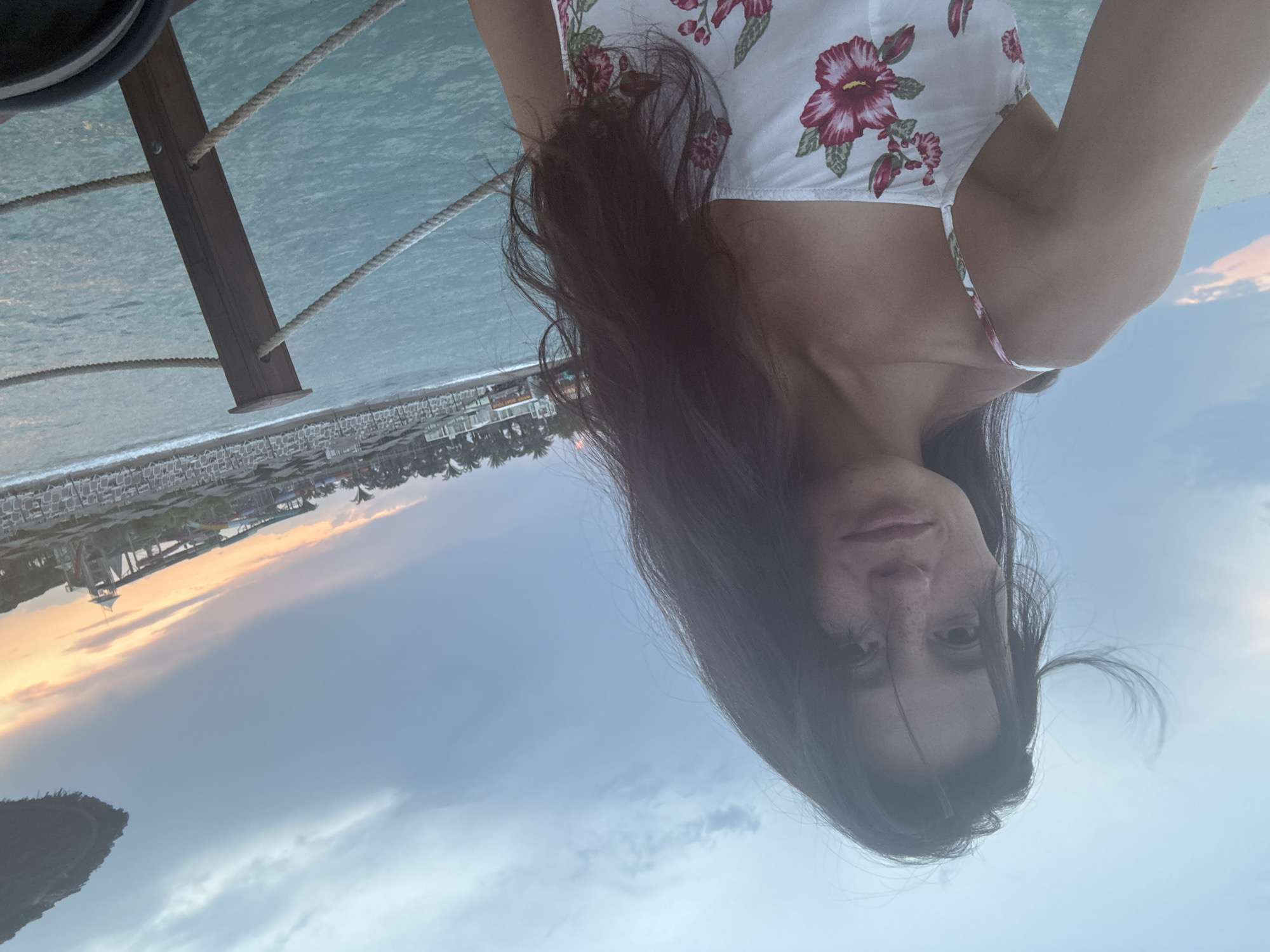 A woman with long, dark hair and freckles on her face takes a selfie from a pier in Aksu, Turkey. She is wearing a white spaghetti strap dress with a floral print. The ocean is calm and blue, with gentle waves. In the background, there is a beach with lounge chairs and umbrellas, and further in the distance, a water park with slides. The sky is cloudy with a hint of sunset colors. A rope railing runs along the pier.