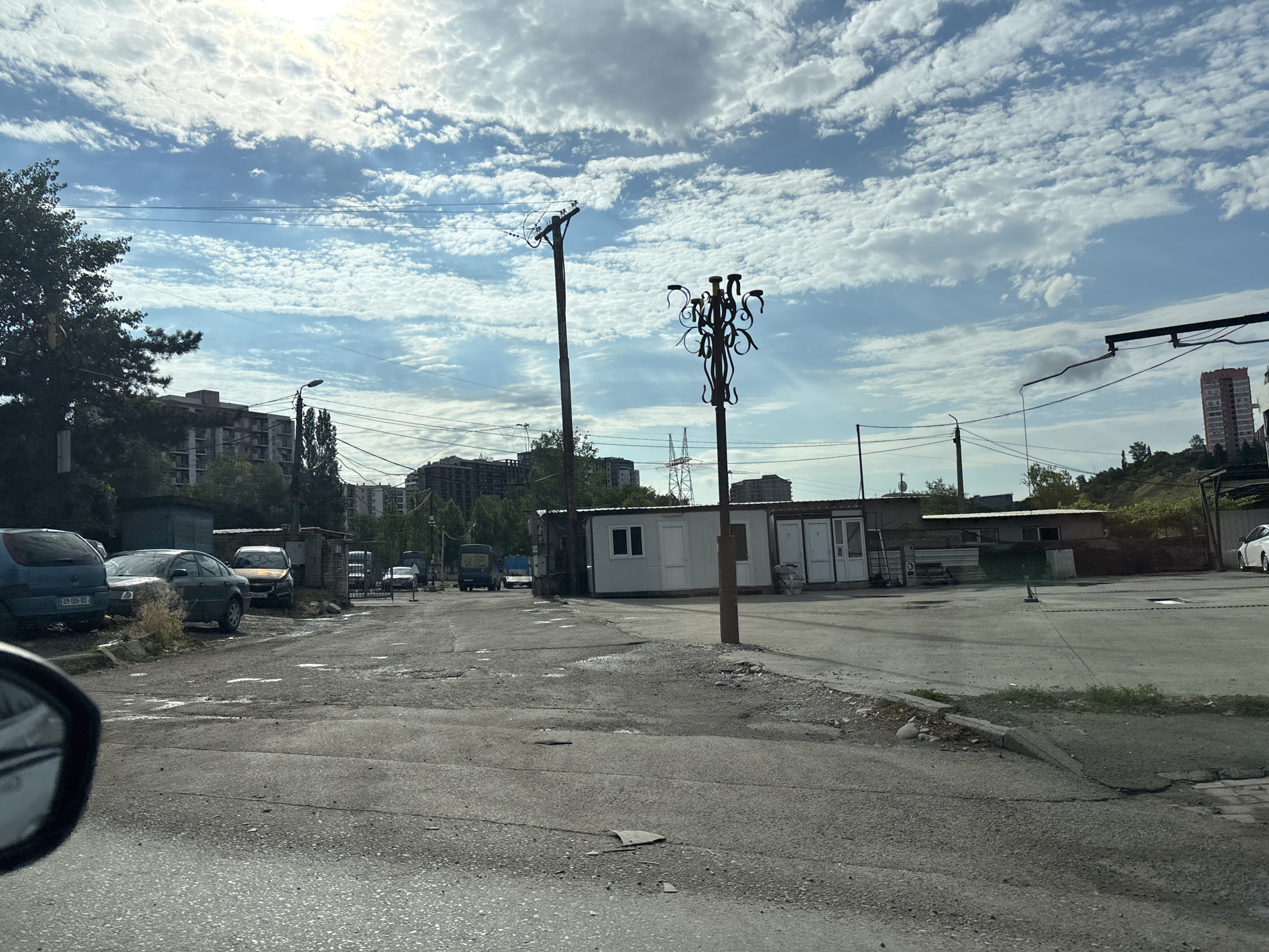 This is a ground-level, exterior, eye-level shot taken from a vehicle, looking towards a paved area with buildings and vehicles. The scene appears to be in Tbilisi, Georgia, as indicated by the license plate on the blue car in the foreground, which reads "AM-006-BO". The sky is partially cloudy with a bright sun.
In the foreground, the asphalt road is rough and uneven, with patches of water or dark stains. A portion of a car's side mirror is visible in the lower left corner. To the left, a blue hatchback car is parked, followed by a dark sedan and another car further down the road. Behind the parked cars, there's a low, dilapidated building or garage.
Further into the scene, on the right side of the road, there's a white modular building with multiple doors and windows, and some sacks of material piled outside. A wooden pole with a decorative metal fixture, possibly an old lamp post without a lamp, stands in front of this building. Several utility poles with wires are also present throughout the area, along with a street light. In the background, apartment buildings and trees can be seen. There are also some industrial-looking structures and power lines.
The area seems to be a parking lot or an industrial/commercial zone, with a mix of old and new structures, and a general sense of disrepair in some parts. No people are visible in the image.