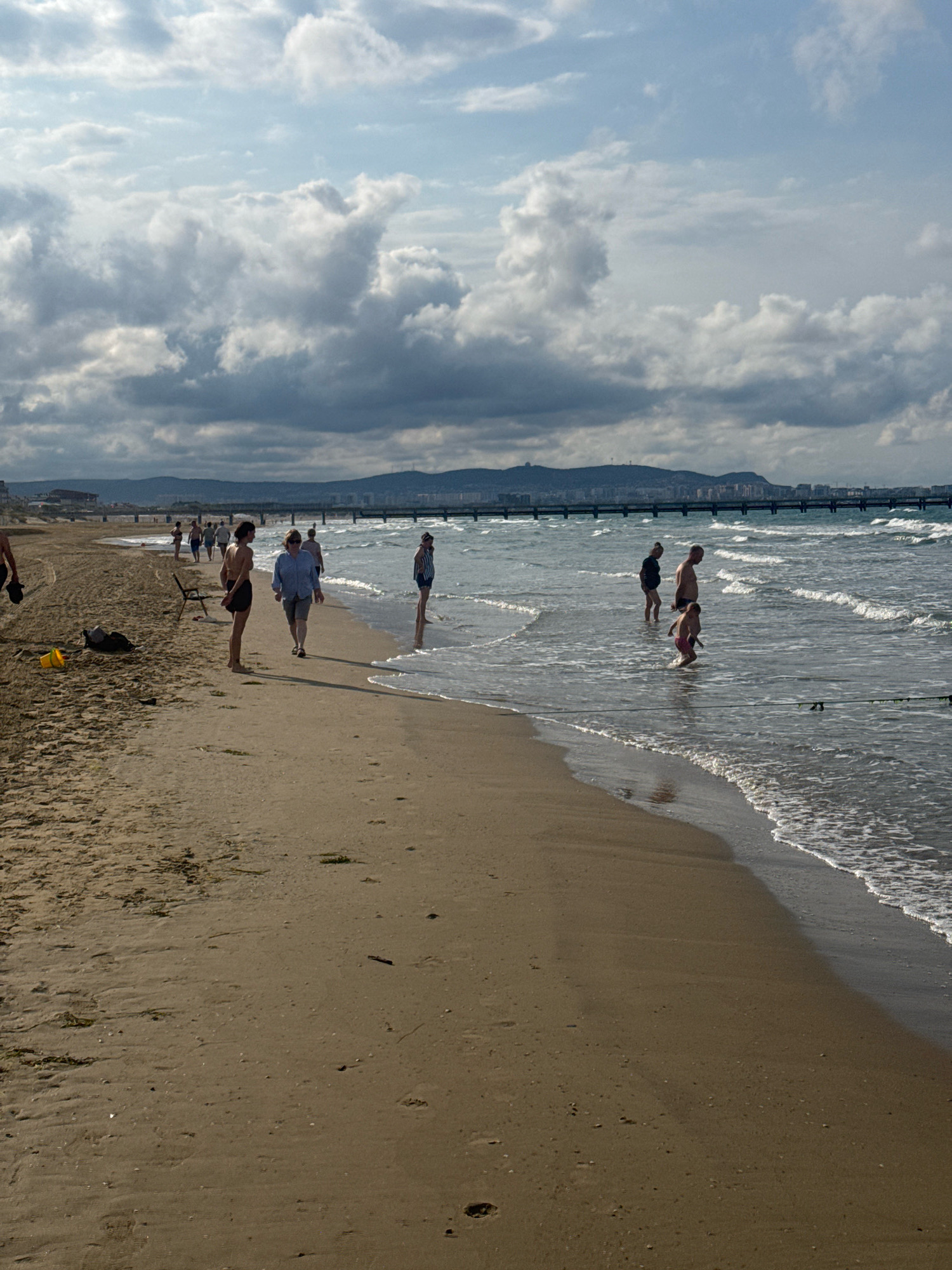 The image shows a wide, sandy beach on the coast of Anapa, Russia. The sea is calm with gentle waves rolling onto the shore. Several people are present on the beach and in the water. In the foreground, a yellow bucket and a dark bag lie on the sand. Further down the beach, people are walking, standing, and wading in the shallow water. In the background, a long pier or bridge stretches across the sea, and a distant city or town is visible on the coastline, backed by rolling hills. The sky is partly cloudy with dramatic, textured clouds, suggesting a mix of sun and shade.