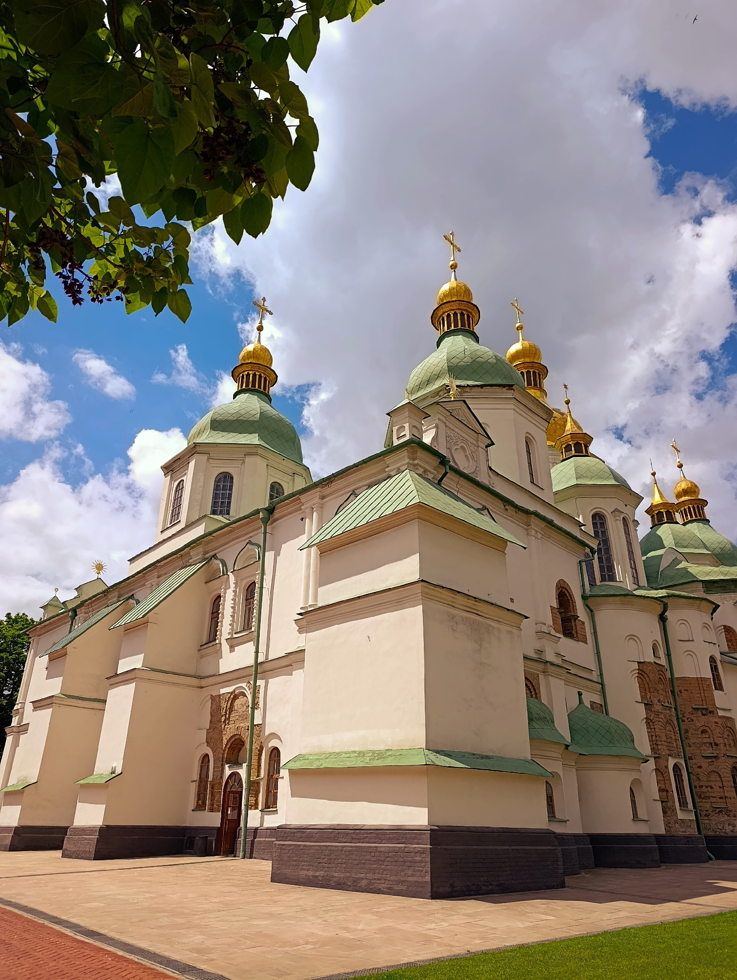 The image captures a low-angle, wide shot of Saint Sophia's Cathedral in Kyiv, Ukraine, under a partially cloudy sky. The architectural marvel features a prominent main building with multiple green-domed towers, each topped with a golden cross. The walls of the cathedral are painted a creamy white, with dark brick detailing at the base. The foreground shows a paved courtyard with sections of light brown paving stones and a strip of green grass to the right. A vibrant green tree with large leaves and some dark seed pods is visible in the upper left corner, partially obscuring the sky. The sky itself is a mix of bright blue patches and scattered, fluffy white clouds, with a few small, dark shapes that could be distant birds or aircraft. The overall scene is bathed in natural daylight, suggesting a daytime setting, likely during the warmer months due to the lush greenery. There are no people or visible signs of activity in the image.