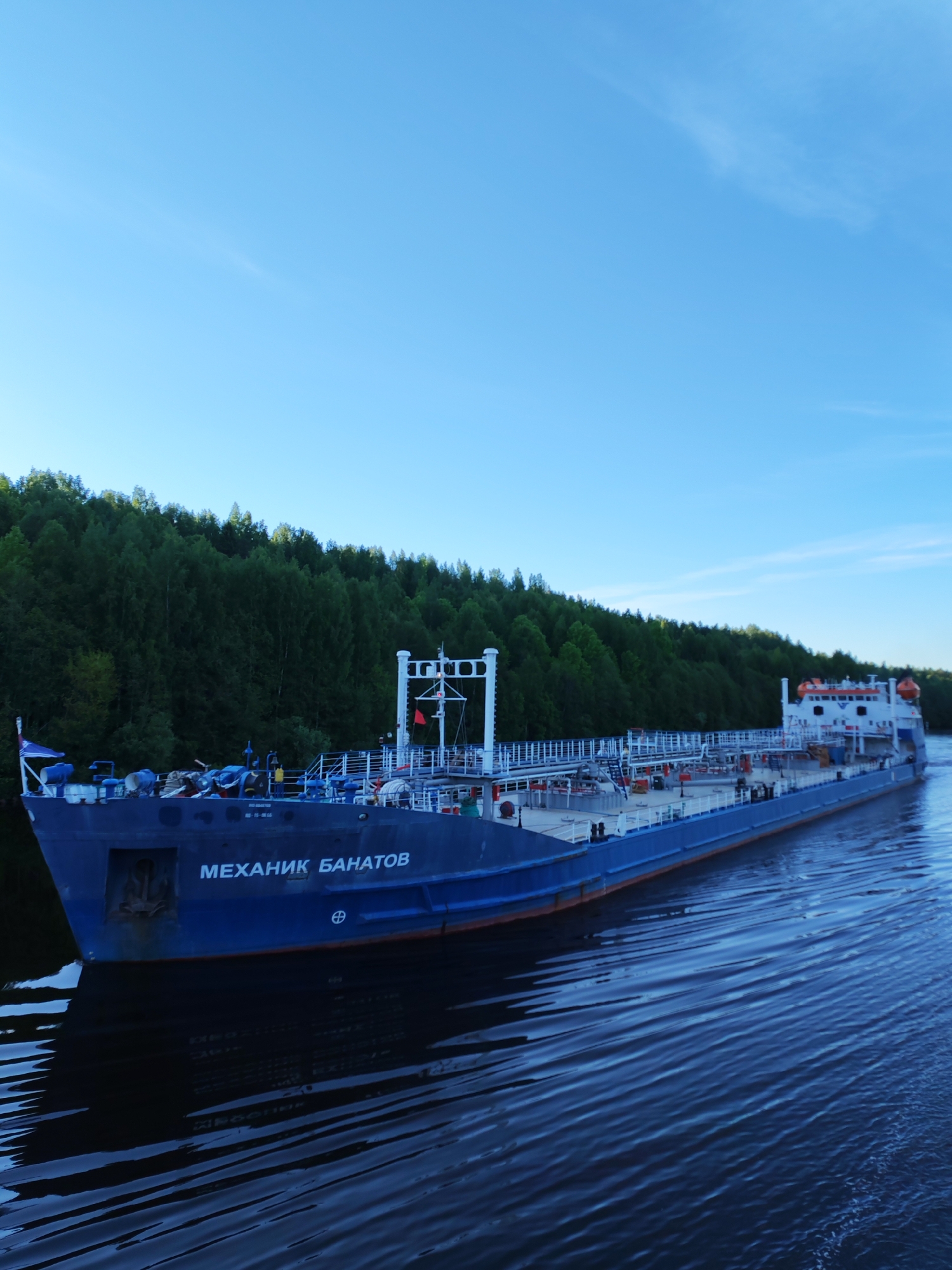 A large blue cargo ship, identified by the Cyrillic text "МЕХАНИК БАНАТОВ" (MEKHANIK BANATOV) on its bow, is shown cruising on a calm body of water in Старое Петровское, Russia. The ship features a prominent blue hull, a white superstructure at the stern, and a deck equipped with numerous pipes, railings, and what appear to be cargo tanks. An anchor is visible on the bow. Orange life rafts are mounted on the ship's stern section. The vessel’s reflection is clearly visible in the dark water, which shows subtle ripples indicating movement.
Along the left bank of the waterway, a dense line of lush green forest creates a natural backdrop. The sky above is a clear, light blue with minimal, faint wispy clouds, suggesting fair weather. The gentle lighting implies either late afternoon or early morning. Another piece of text visible on the ship, smaller and above the main name, reads "НЕ-15-08.58". The overall scene conveys a sense of tranquility as the ship navigates the peaceful waters.
