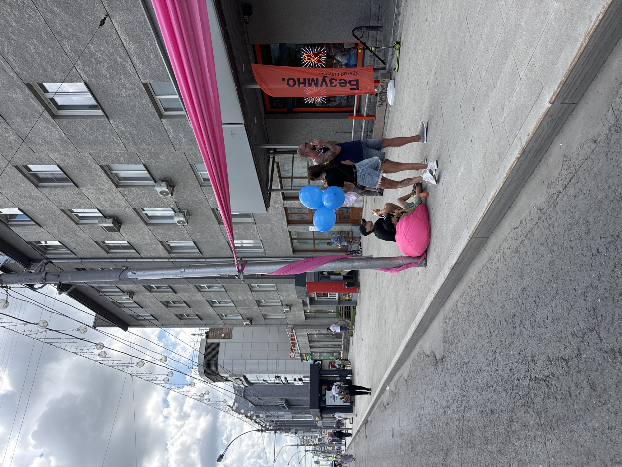 A street scene in Novosibirsk, Russia, captures a sidewalk adjacent to a gray, multi-story building. String lights with circular bulbs are strung across the street. A large, bright pink fabric is draped diagonally across the building's facade. On the sidewalk, a group of people are gathered. In the foreground, two individuals sit on a large, pink beanbag chair, one with a drink in hand. A man stands nearby holding a cluster of four blue balloons. To his right, a tall, orange flag with Cyrillic text, "Безумно. Крутая шаурма." which translates to "Crazy. Cool Shawarma," is planted in a white base. A green scooter is parked on the sidewalk. In the background, other pedestrians are visible walking on the sidewalk and street. The sky is overcast with clouds.