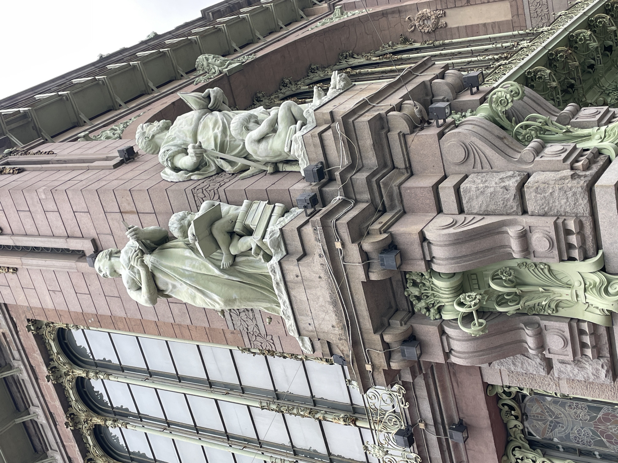 A close-up view of the Singer House building in Saint Petersburg, Russia. The building's facade features statues depicting a woman and a child. The sculptures are positioned above a large window and set against the textured brickwork of the building. Below the statues, intricate ornamental details are visible. Green embellishments are integrated into the design, and the window frames are decorated with scrollwork. Small spotlights are attached to the building's structure, and the overall aesthetic emphasizes a blend of architectural elements and sculptural artistry.