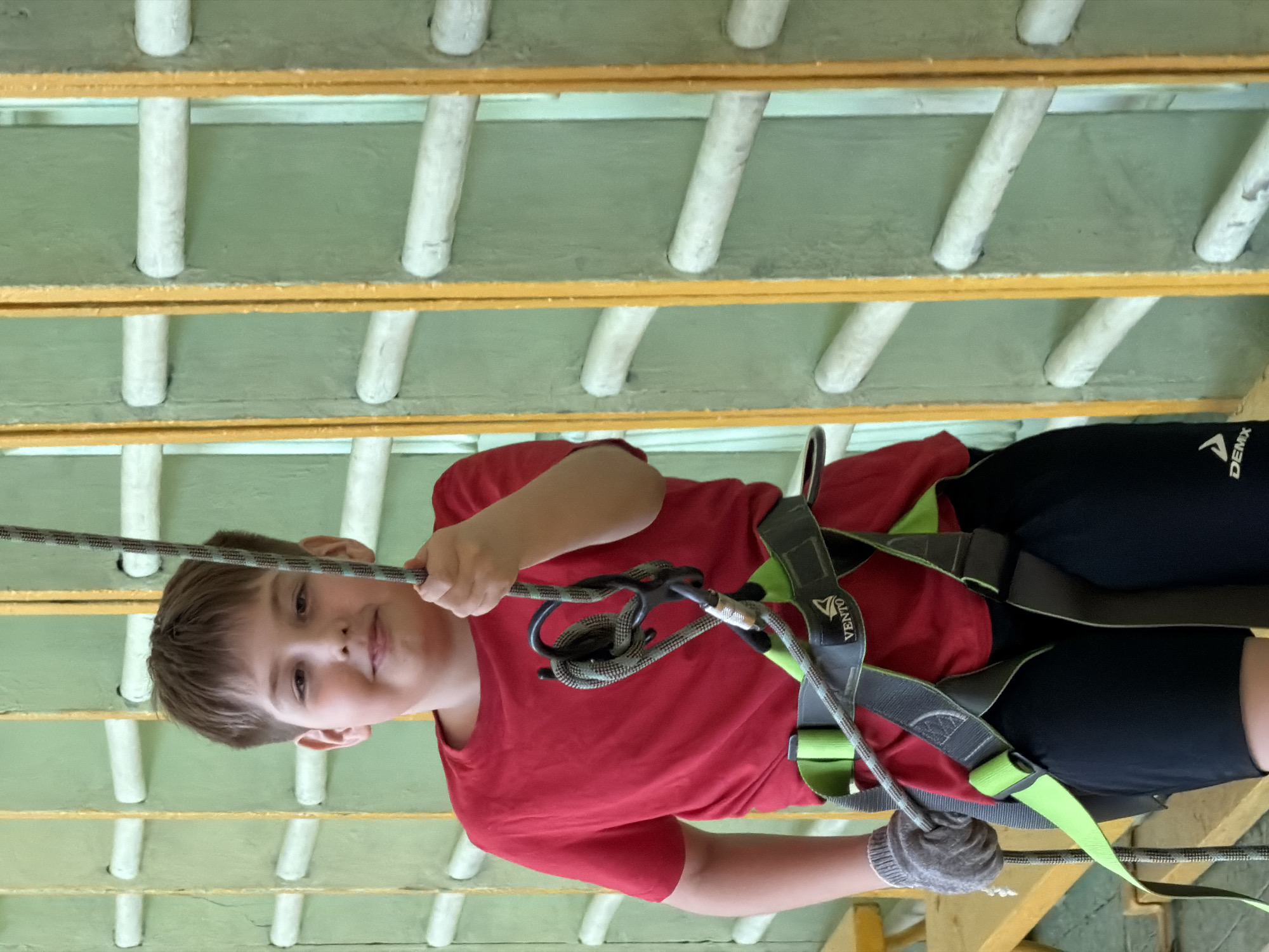 A young boy is shown suspended in the air, wearing a safety harness and gloves, grasping a rope. The harness is gray with green accents. He is wearing a red t-shirt and black shorts. The background consists of wooden beams and cylindrical supports, painted white. The location is Izhevsk, Russia.
