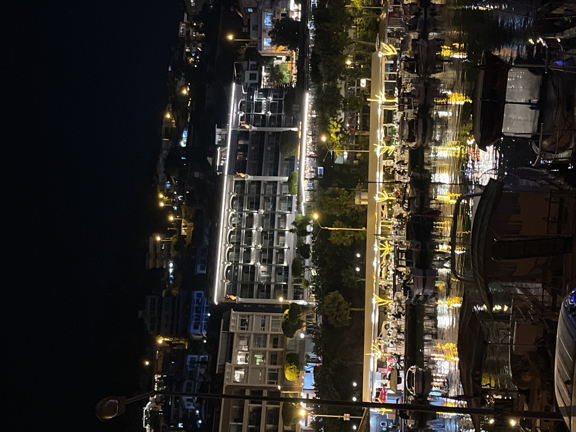 This image captures a vibrant night scene in Alanya, Turkey, showcasing a lively waterfront area. The setting is dark, illuminated by numerous artificial lights from buildings, streetlights, and decorative features, casting a warm glow over the entire scene.
On the left, a series of multi-story buildings, likely residential or hotels, line a distant hillside. These buildings are well-lit, with many windows showing interior lights and some featuring exterior architectural lighting, creating a bright skyline against the dark sky.
In the mid-ground, a bustling promenade stretches horizontally, separating the buildings from the water. It's adorned with trees and streetlights, and structures along this strip are brightly lit, including what appear to be restaurants or cafes with outdoor seating. Decorative lights, shaped like arrows or stars, are prominently displayed along the promenade's edge, adding to the festive atmosphere. People are visible sitting at tables, suggesting dining or socializing activity.
To the right, a body of water, likely a harbor or canal, is filled with docked boats of various sizes. The most striking feature of this area is the intense reflection of all the surrounding lights on the water's surface. These reflections create shimmering patterns of yellow, white, and other colors, giving the water a dynamic and almost abstract appearance. The reflections are so strong that they obscure clear views of the water itself in many places.
The overall impression is one of a busy and appealing evening destination, with a focus on nightlife and leisure by the water. There is no visible text that is clearly legible within the image.