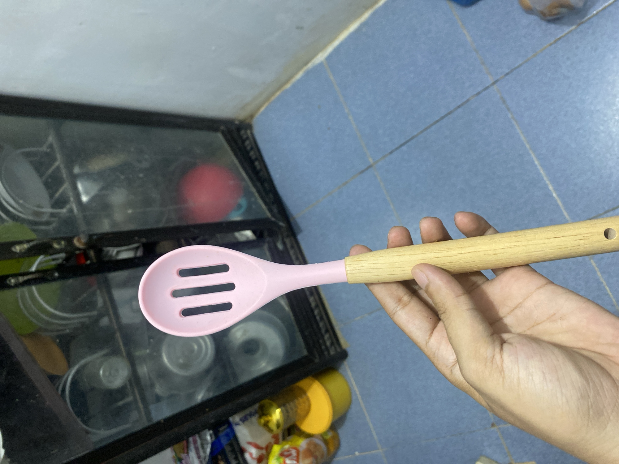A close-up, eye-level shot shows a person's hand holding a pink silicone slotted spoon with a light-colored wooden handle. The spoon is positioned vertically, with the head of the spoon filling the upper portion of the frame. The hand is light-skinned and appears to be gently gripping the wooden handle. Behind the hand and spoon, a glass-fronted cabinet filled with various kitchen items, including plates and containers, is visible. To the left of the cabinet, a bright yellow plastic container is partially visible. The floor is covered in blue square tiles with white grout lines. The scene appears to be an indoor domestic environment, possibly a kitchen, located in the city of Lhokseumawe, Indonesia.