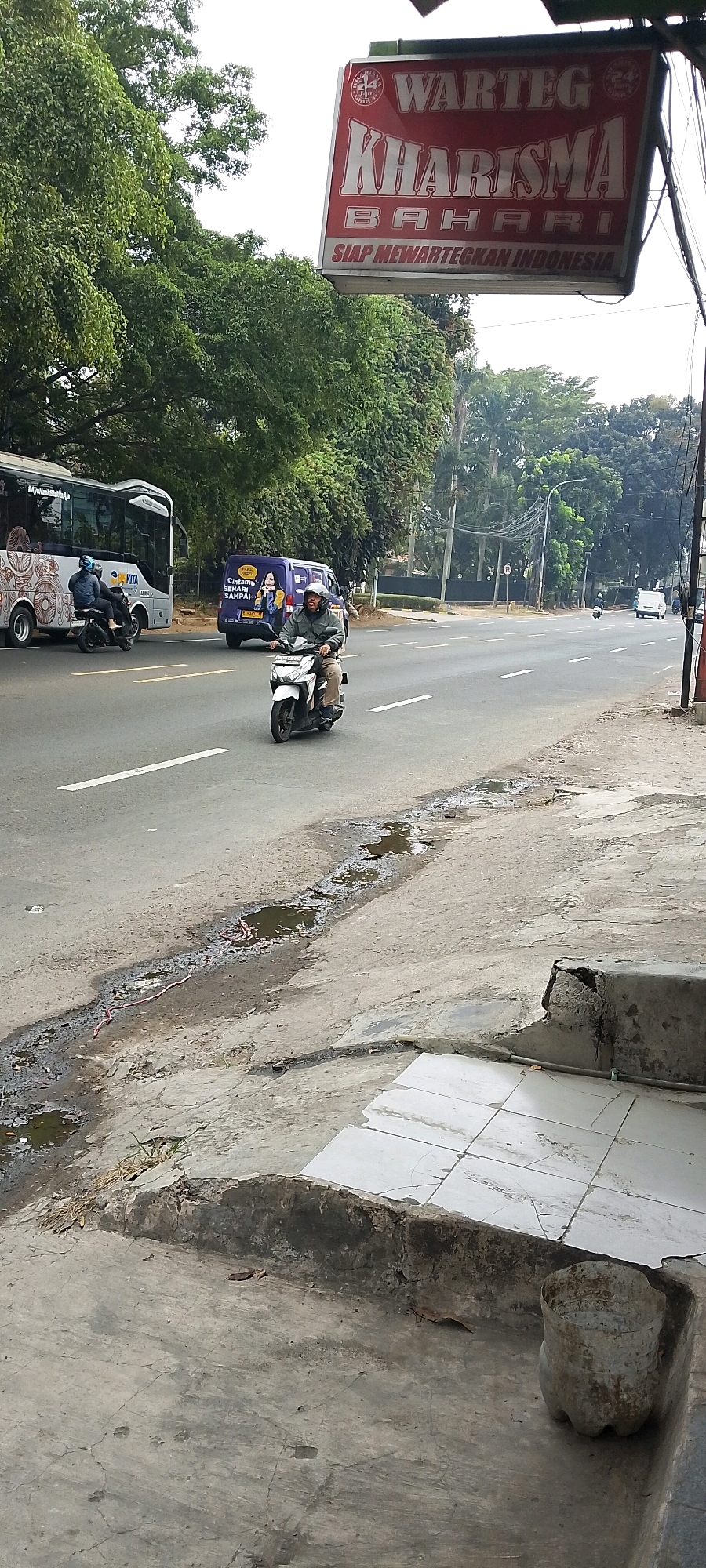 The image shows a street scene in Depok, Indonesia, near a restaurant called "Warung Kharisma Bahari". On the road, a person is riding a white motorcycle. To the left, a bus is visible with passengers on board, and a purple van is parked. Trees line the street, and a sign for the restaurant is prominently displayed. The foreground of the image shows a cracked concrete sidewalk with some puddles and debris, suggesting an outdoor environment in a city.