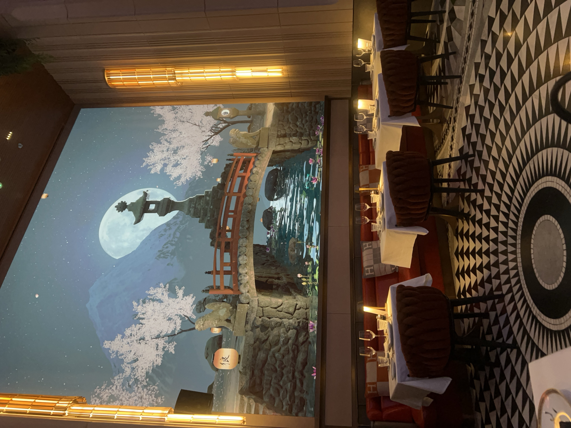 This image captures the interior of an upscale restaurant, likely in Dubai, UAE, characterized by its distinctive decor. The main focal point is a large, immersive mural or projection on one wall, depicting a serene, stylized Japanese landscape at night.
The mural showcases a massive full moon illuminating a mountainous backdrop, possibly inspired by Mount Fuji, under a starry sky. Cherry blossom trees with white flowers are prominent in the foreground and midground. A traditional red arched bridge spans over a body of water, which reflects the surrounding scenery and features lily pads with small pink flowers. Stone lanterns are placed throughout the landscape, with one notably displaying Japanese characters. A stone statue, resembling a guardian lion-dog, sits on the bridge structure.
The dining area is arranged with multiple tables, each set with a white tablecloth, several wine glasses, and what appears to be dinnerware. The chairs are upholstered in a dark brown or reddish material with a ribbed texture, suggesting comfort and elegance. Luxurious, warm-toned lighting fixtures are integrated into the ceiling above the mural, and smaller lamps illuminate each table. The floor features an elaborate black and white geometric pattern, with concentric circles and radiating triangular motifs, adding a bold and sophisticated element to the space. Red banquette seating with decorative pillows, some bearing an 'H' pattern, is visible behind some tables.
No people are present in the image, suggesting it was captured during a quiet moment, perhaps before opening or after closing. The warm lighting and detailed mural create an elegant and tranquil atmosphere, ideal for an evening dining experience. The visible Japanese character on the red lantern in the mural appears to be "祝" (shuku), meaning "celebration" or "congratulations." The overall aesthetic points to a high-end establishment with a clear Asian, specifically Japanese, thematic influence.
