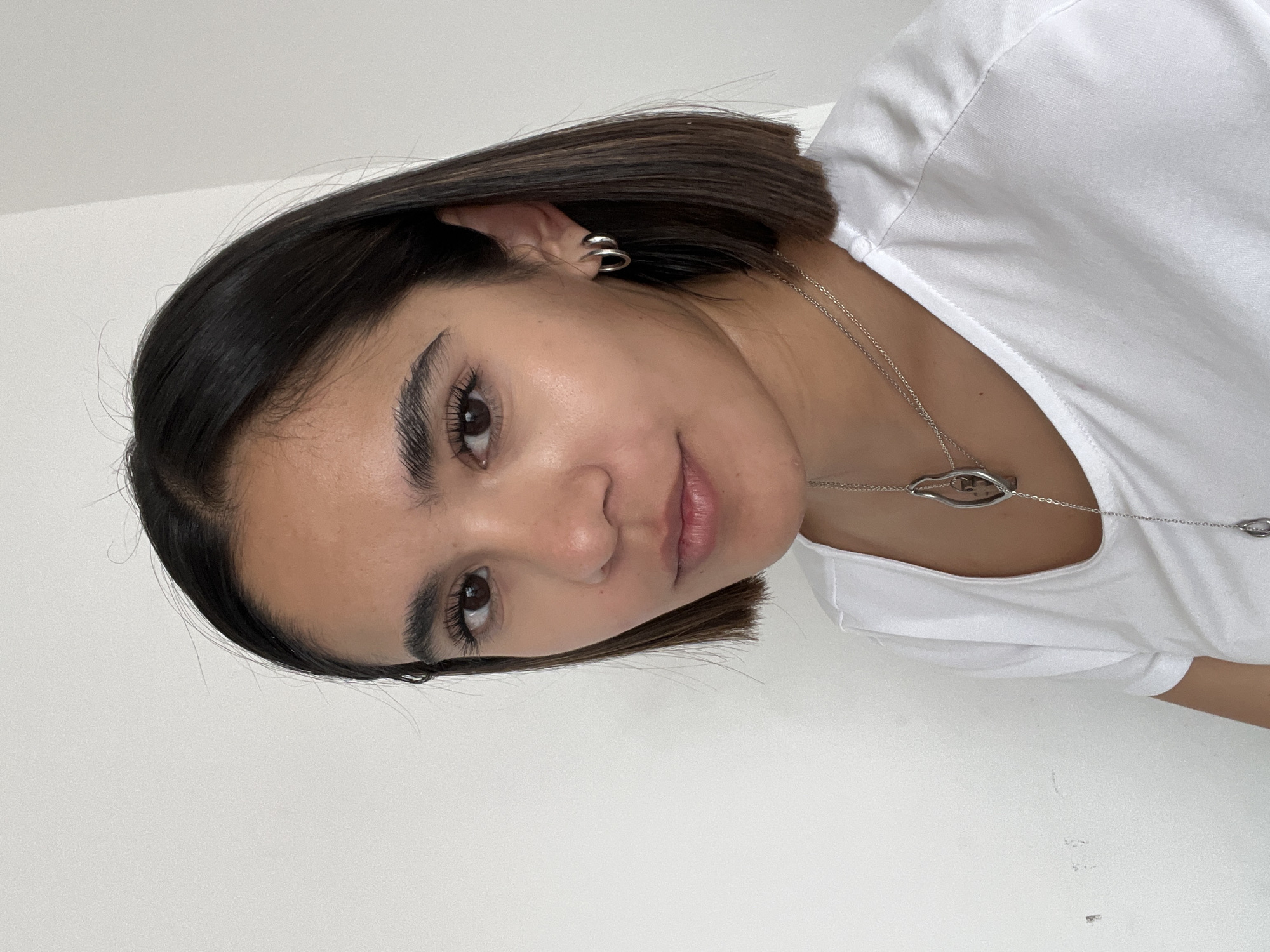 A medium close-up photograph shows a young woman with dark brown hair and fair skin. She is wearing a white t-shirt and a silver necklace. She appears to be indoors against a white wall. The location is not discernible from the image.