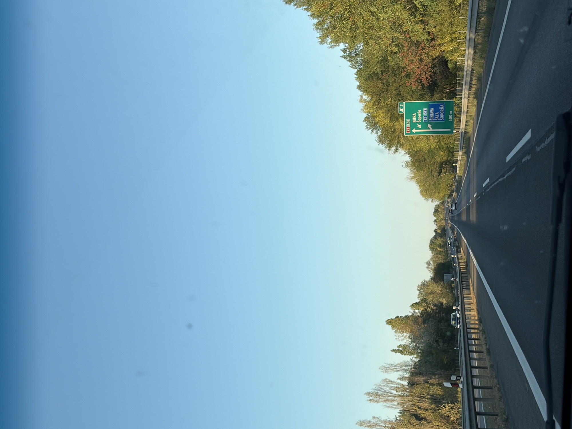 The image shows a view from a vehicle traveling on a highway in Dolná Streda, Slovakia. A green highway sign is visible on the right side of the road, indicating directions to Nitra, Soporna, Sintava, Sala, and Soporna, with a distance of 500 meters. Trees line both sides of the highway, with some showing autumn foliage. The sky is clear and blue. The road has multiple lanes, and there are other vehicles on the highway, including cars and trucks. A metal guardrail separates the lanes. The scene appears to be captured during daylight, likely in the late afternoon, given the lighting conditions.