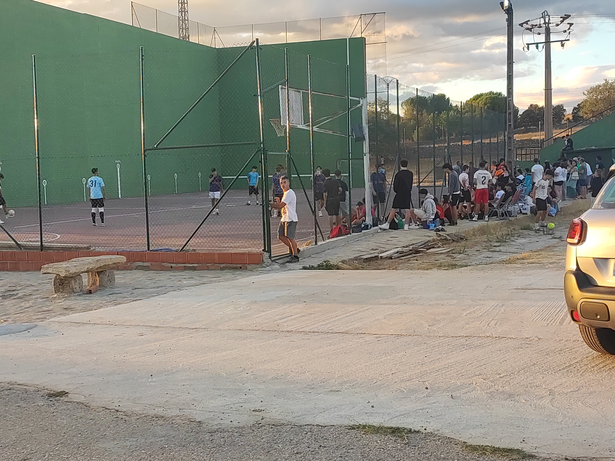 The image depicts an outdoor sports facility, likely in Sobradillo, Spain, with a large green wall that serves as a backdrop for a game. Several individuals are present, some actively participating in a sport on the court and others gathered outside the fenced area. A basketball hoop is visible on the green wall. The ground is a mixture of sand and paved surfaces. A stone bench is situated on the sand to the left of the frame. In the background, trees and power lines suggest a semi-urban or rural setting. The lighting indicates it is late afternoon or early evening, with the sun casting long shadows. A silver car is parked on the right side of the image.