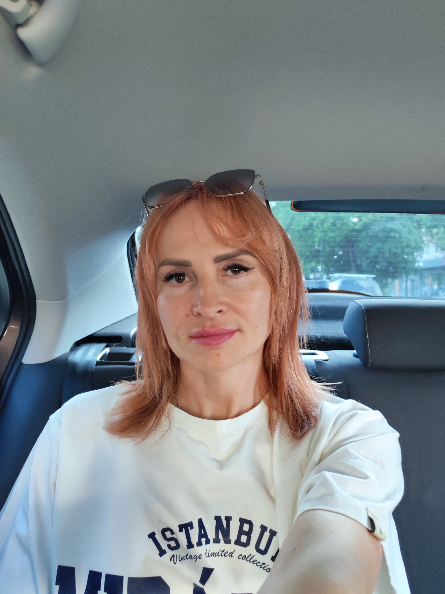 A woman with reddish-blonde hair and sunglasses perched on her head is taking a selfie from the back seat of a car. She is wearing a white t-shirt with dark blue text that reads "ISTANBUL Vintage limited collection". The background shows the interior of a car, including the headrests and a portion of the ceiling. Through the car window, trees and buildings are visible, suggesting an outdoor setting. The lighting appears to be natural daylight, indicating it is daytime. The woman's expression is neutral. The location is Chaikovsky, Russia.