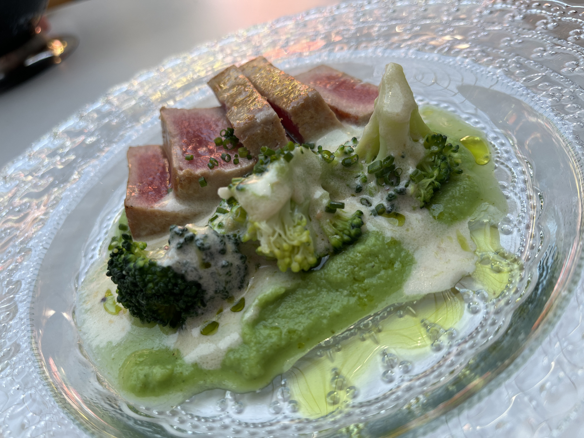 A close-up, overhead shot displays a gourmet dish of sliced seared tuna and broccoli florets served on a clear, textured glass plate. The tuna, cut into thick, rectangular pieces, exhibits a pink, seared exterior and a rare, reddish interior. It is arranged in a row, drizzled with a creamy white sauce and garnished with finely chopped chives. The broccoli is presented in clusters, interspersed with the tuna, and also covered in the creamy sauce and a vibrant green purée. A drizzle of olive oil is visible on the green purée and scattered across the plate. The plate itself has a decorative, raised patterned rim and a glossy finish, with subtle reflections of light and color. The background is blurred, suggesting an indoor setting with warm, ambient lighting. The overall impression is of an elegantly plated, sophisticated dish.