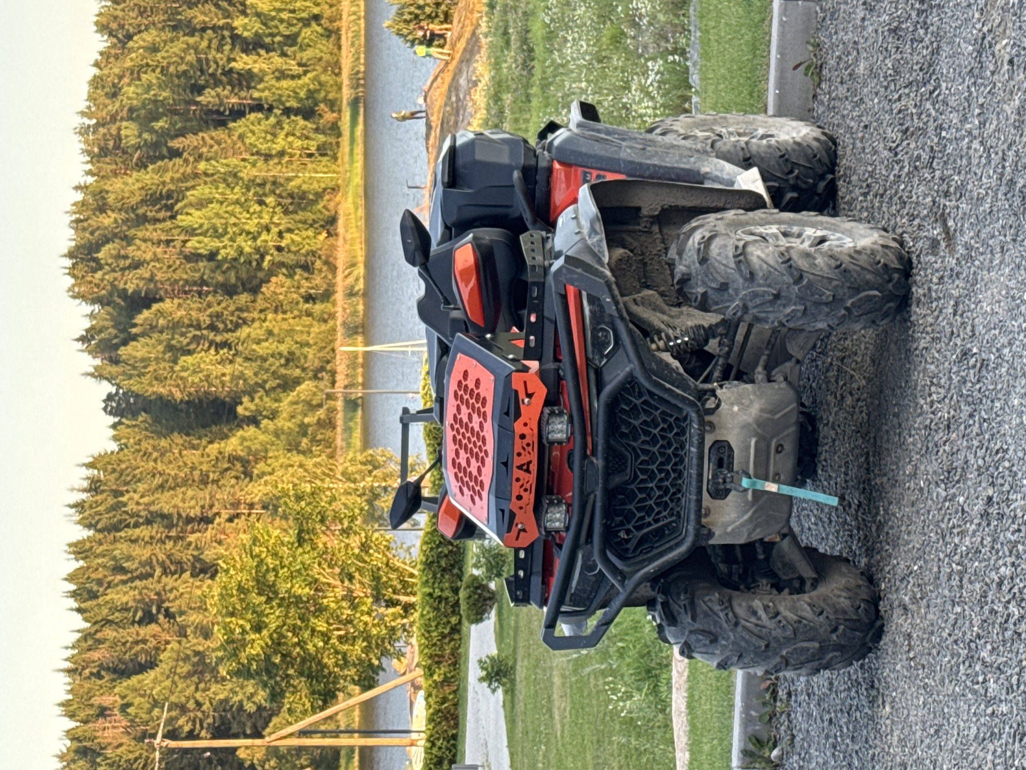 An all-terrain vehicle (ATV) is parked on a gravel surface in front of a body of water, possibly a lake or a river, with a dense forest in the background. The ATV is red and black, with large, knobby tires. It has a winch visible on the front and two headlights. The surroundings include green grass, some low bushes, and wildflowers. In the distance, on the opposite bank of the water, several people are visible, some standing near the water's edge and others further back. The sky is clear and appears to be late afternoon or early evening, with the sun casting a warm glow on the trees. The location is identified as Kurkijoki, Russia.