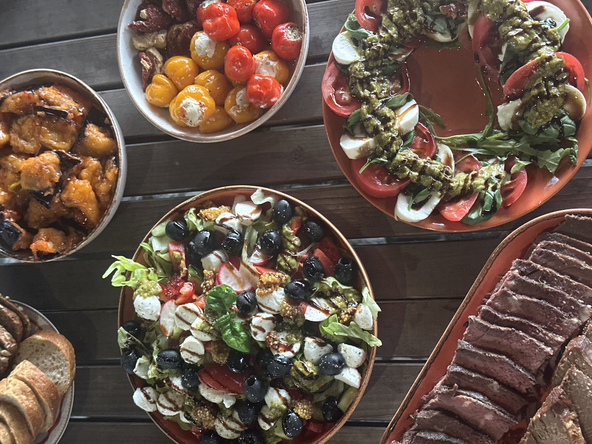 A top-down shot displays a wooden table laden with various food dishes. A bowl of glazed eggplant sits in the top left, near a bowl of stuffed peppers. Above the center, a tomato and mozzarella salad is arranged in a ring on an orange plate. Below the center, a mixed salad with mozzarella balls and olives is displayed. To the bottom left is a plate of sliced bread, and to the bottom right is a platter of sliced meat. The setting appears to be a restaurant or private event, but there is no clear indication of a location like Aviator, Russia. There are no people visible in the frame.