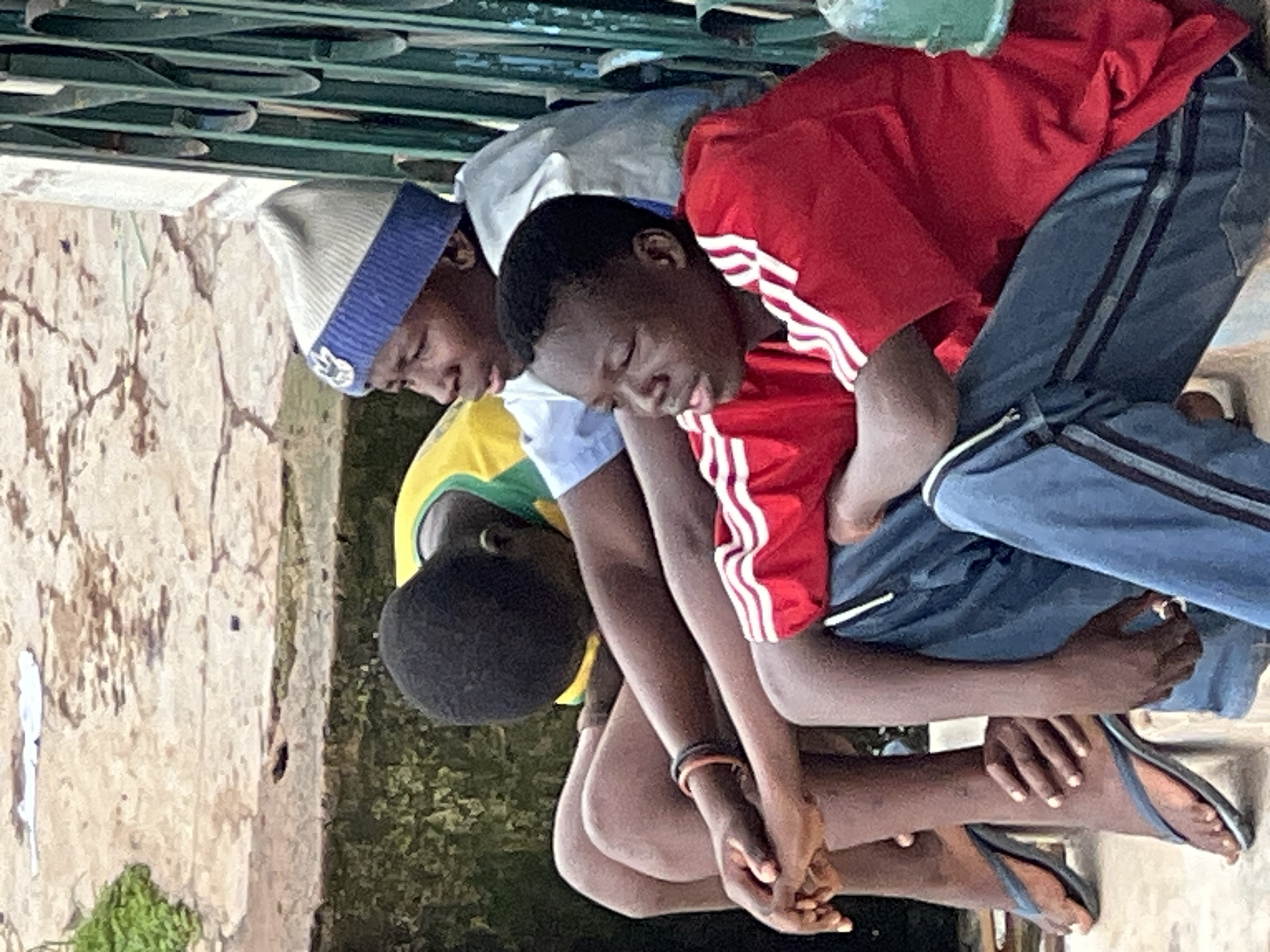 Three young males are seated next to each other on a concrete ledge in a seemingly urban environment. The individual on the far left wears a yellow and green shirt and has his head bowed, obscuring his face. The middle subject is wearing a blue and white knitted beanie and a light blue shirt, with his face turned to the left, looking away from the camera. The individual on the right is wearing a red t-shirt with white stripes on the sleeves and dark blue pants. His face is visible, looking to the left. Their legs are crossed and folded in front of them. The environment appears somewhat weathered, with cracked concrete and some greenery in the background. A green metal railing is visible to the right of the frame. The lighting suggests it is daytime, possibly overcast. The scene does not provide specific visual cues to pinpoint the exact location within Jos, Nigeria, beyond its general urban characteristics.