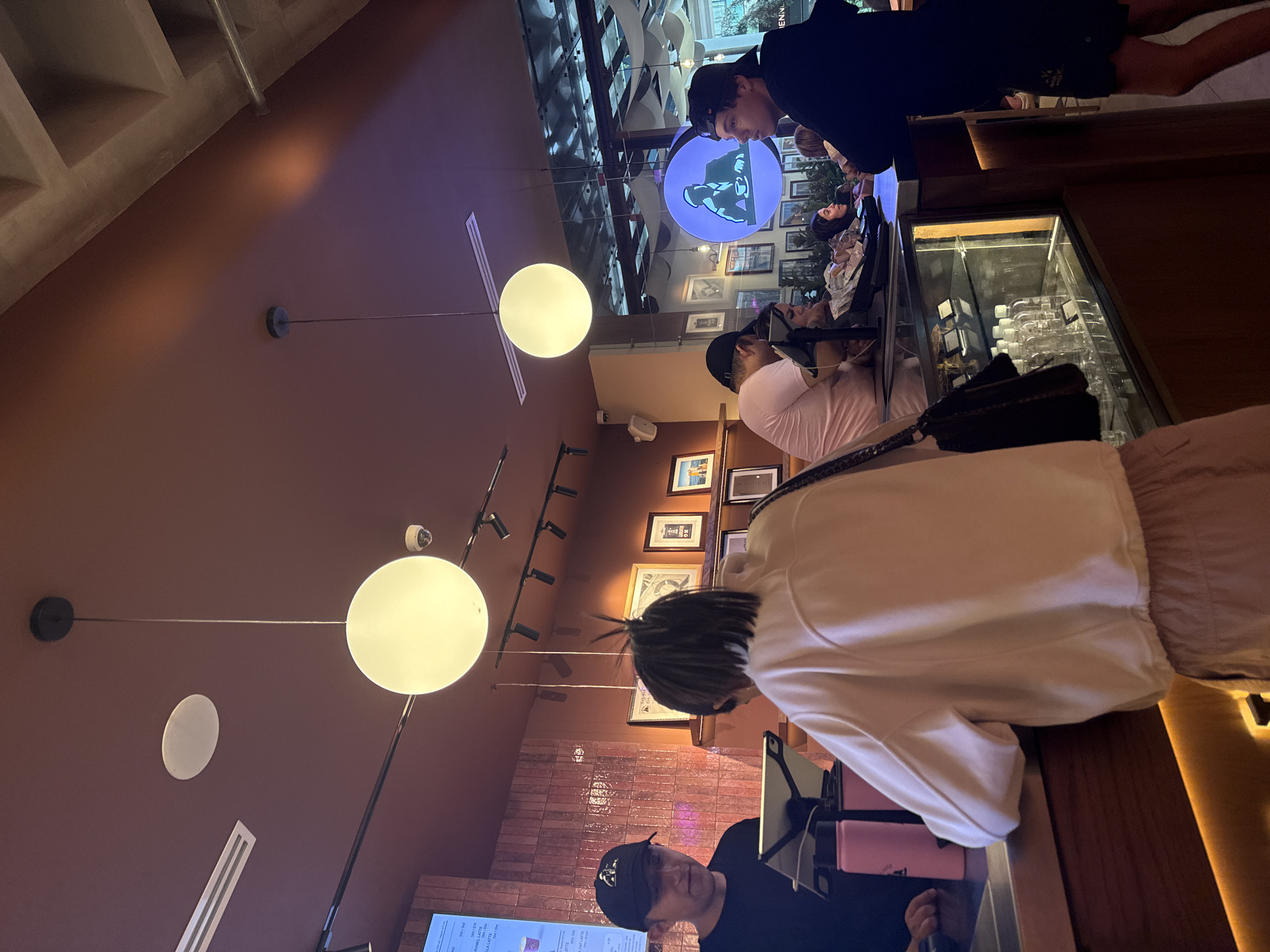 Here is a description of the image:
The image shows the interior of a café or restaurant in Mexico City, Mexico. Several people are present, some standing at the counter to order or pay, and others seated at tables. The scene is illuminated by modern globe light fixtures hanging from the ceiling. The walls are a dark reddish-brown color, with framed pictures lining the walls further back in the space. There is a display case behind the counter filled with glassware or other items. The counter has a pink device. The overall atmosphere appears to be casual and bustling with activity.