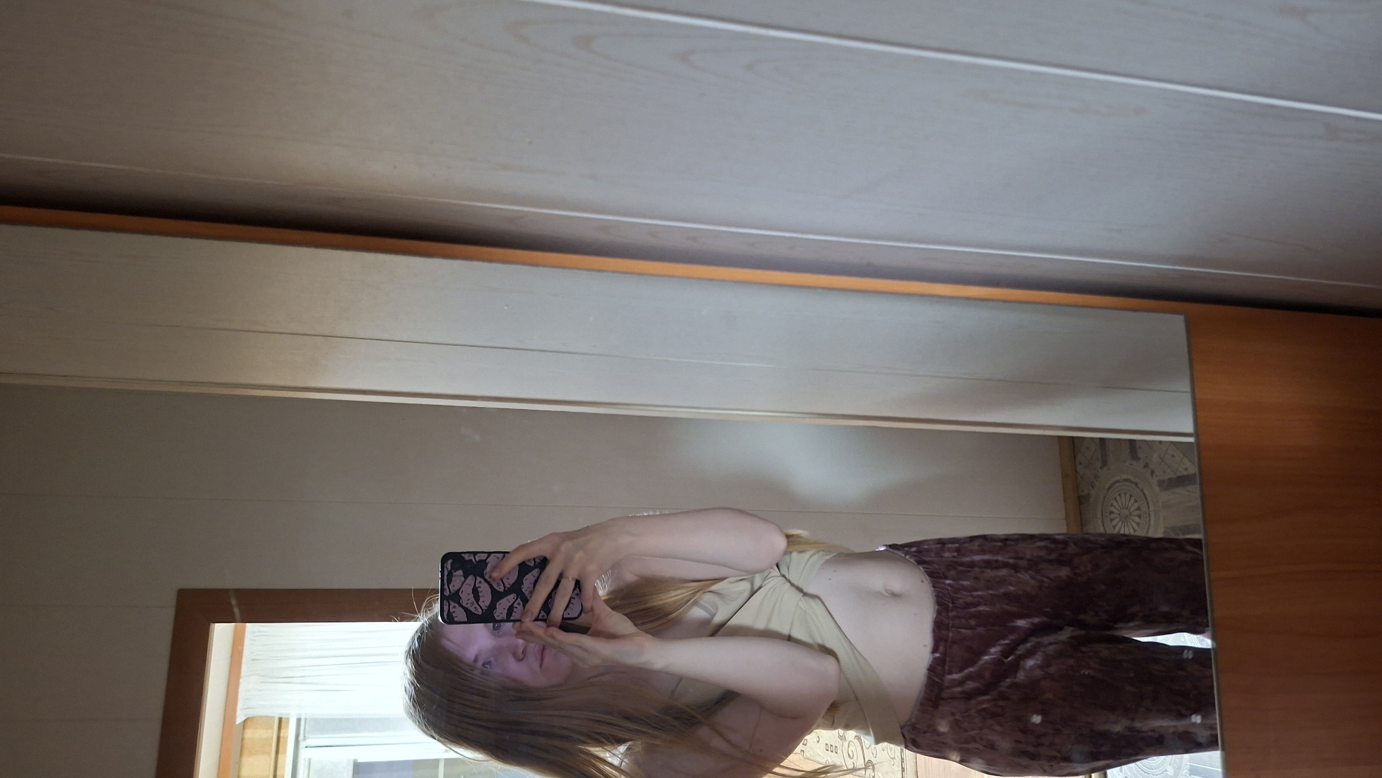 A mirror selfie shows a young woman with long blonde hair, wearing a tan top and maroon velvet pants. She is holding a smartphone with a black and pink patterned case in front of her face. The mirror reflects a room interior with light-colored wood paneling on the walls and ceiling. A window with sheer white curtains is visible in the reflection. Part of a patterned floor can also be seen in the reflection. There is nothing to suggest that this photo was taken in Большой Букор, Russia.