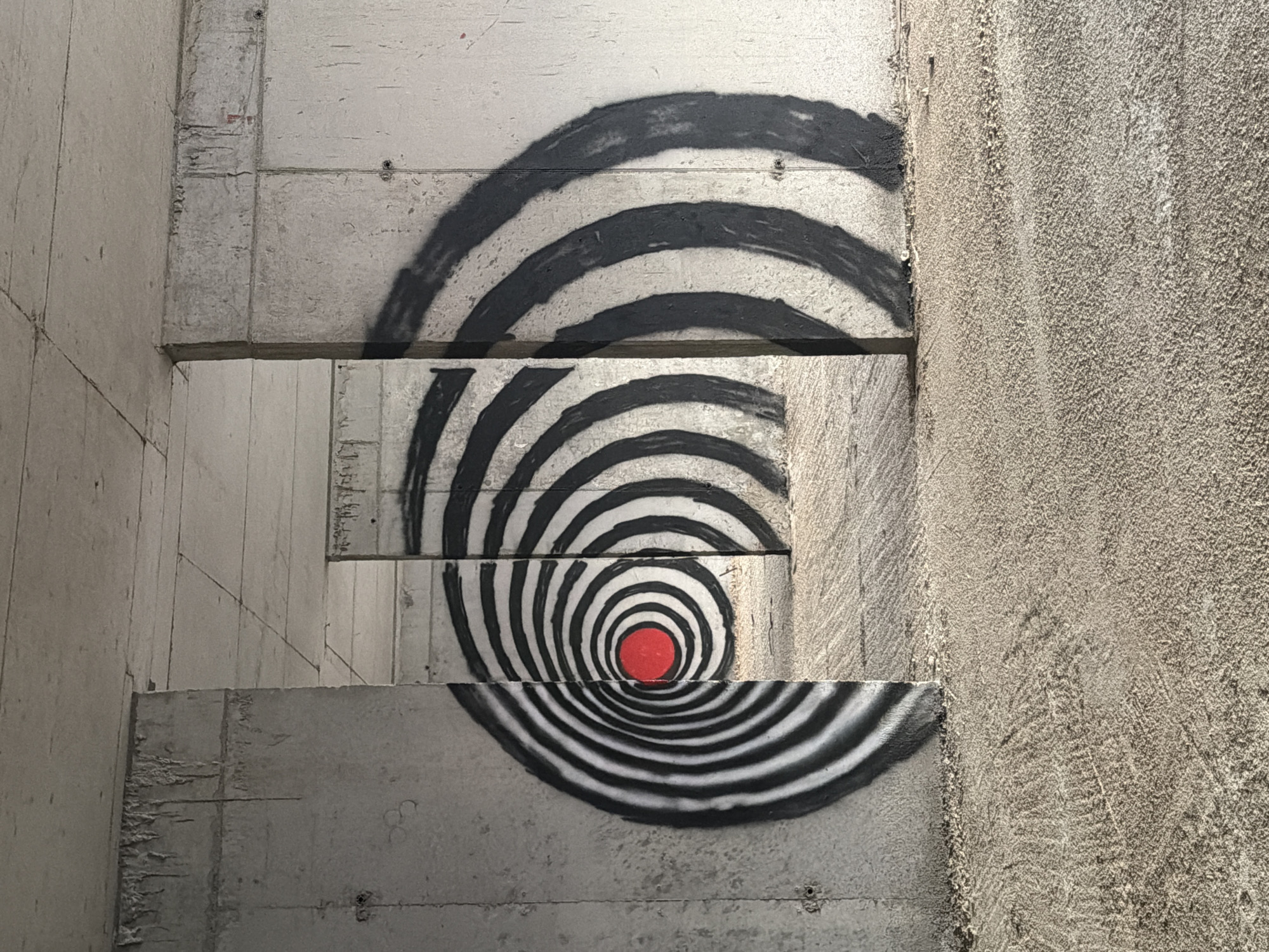 An outdoor shot captures graffiti on a concrete structure, possibly an underpass or a part of a construction site, in Perazića Do, Montenegro. The graffiti features concentric circles in black and white, with a bright red circle at the center of one of the spirals, creating a target-like or hypnotic effect. The concrete walls are rough and unfinished, showing signs of wear and tear. The ground is covered in loose gravel and sand. The perspective suggests a tunnel-like environment, with openings leading further into the structure. The lighting indicates daytime, with shadows cast by the graffiti and the architectural elements. There are no people or animals present in the scene.