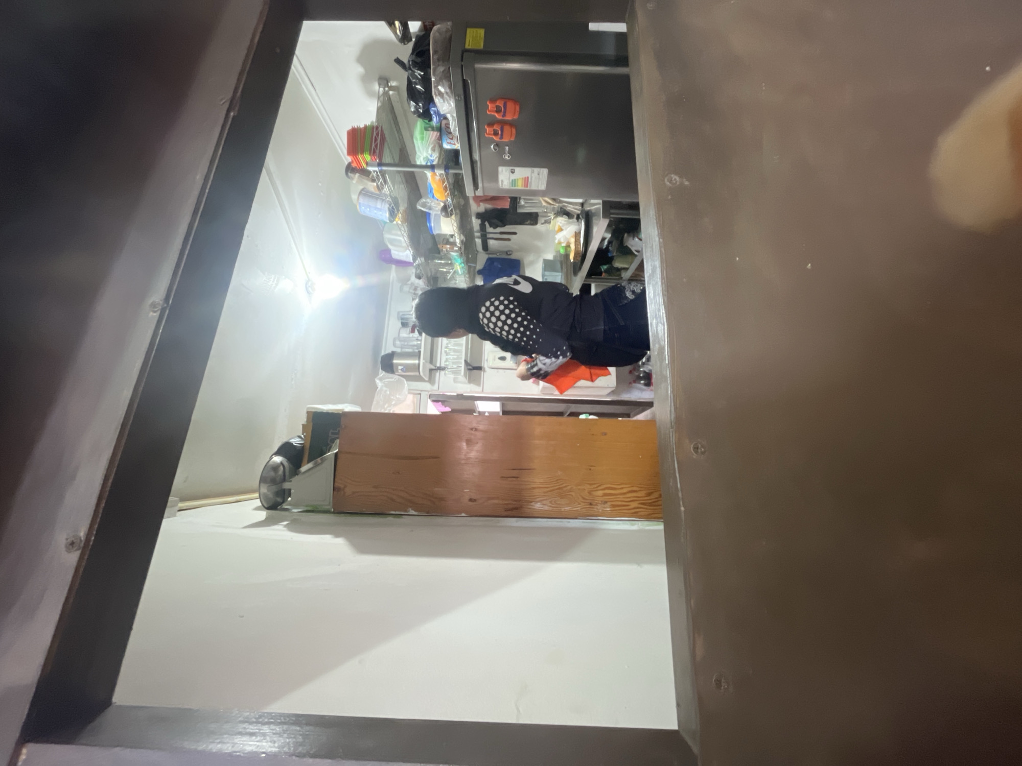 The image depicts an indoor scene, possibly a kitchen area, viewed through a dark wooden frame. A person, wearing a dark vest with white polka dots over a long-sleeved shirt and dark pants, is standing in front of a stainless steel refrigerator. They are holding an orange cloth in their left hand and appear to be in the process of cleaning or organizing. The kitchen is equipped with shelves stocked with various containers, plates, and kitchen utensils. A tall, wooden cabinet stands to the left of the person. The overall lighting suggests an artificial light source is illuminating the space. The scene is consistent with a commercial kitchen or a domestic kitchen in a densely populated area. Given the specific details of the image, it is not possible to definitively determine the exact location within Iquique, Chile, without additional contextual information or external references.