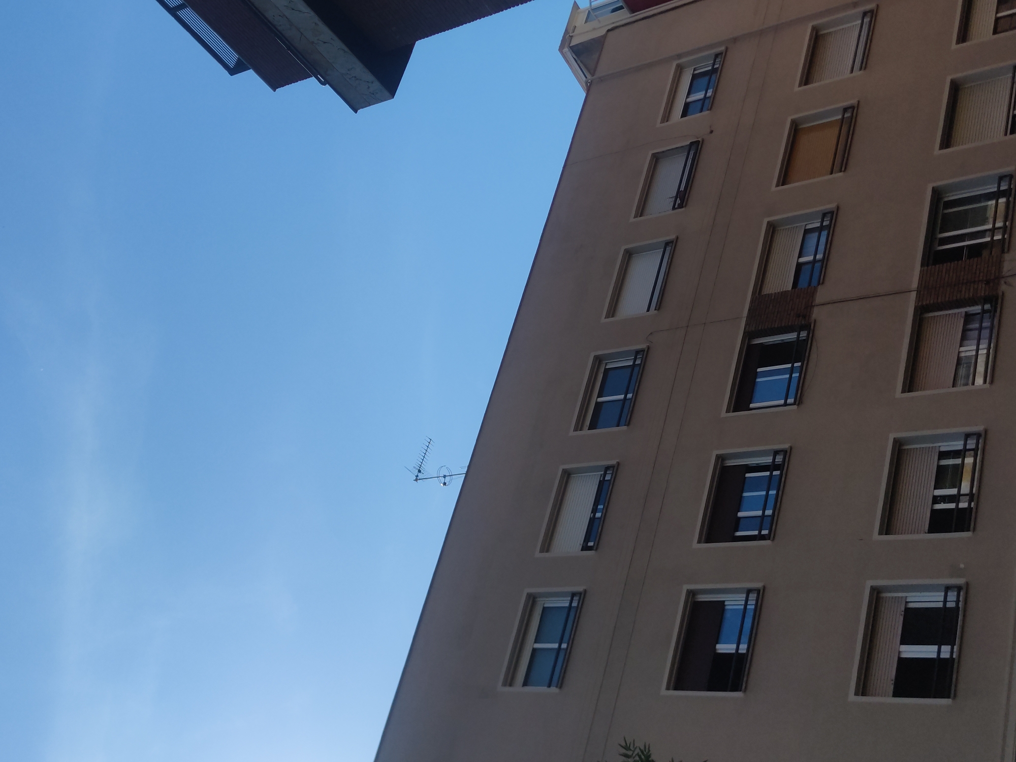 The low-angle shot captures a tall beige building, seemingly an apartment block, juxtaposed against a clear blue sky. The building's facade is punctuated by a grid of windows, some fitted with open metal screens, allowing glimpses of the interior. An old-fashioned television antenna is attached to the side of the building. Another building is present in the top left of the frame. The photograph appears to be taken in an urban environment.