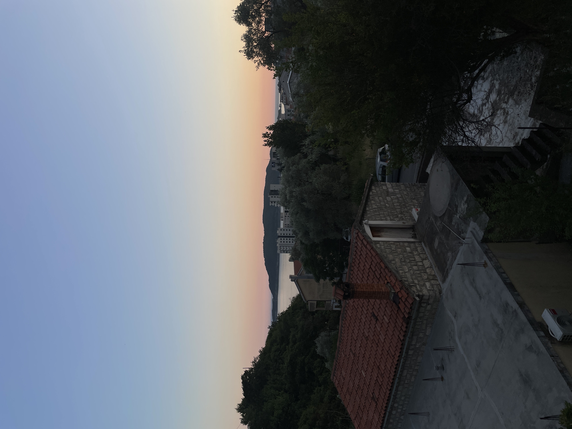 The image depicts a dusk scene over the town of Rafailovići, Montenegro, with the Adriatic Sea visible in the distance. In the foreground, a building with a red-tiled roof and stone walls is prominent. The roof is flat and appears to be under construction or maintenance, with several metal rods protruding from it. Adjacent to this building is another structure with a similar stone facade and a wooden door.
The middle ground features a dense canopy of green trees, with some olive trees interspersed. Beyond the trees, several multi-story buildings, likely apartments or hotels, are visible, some with modern architecture and balconies. A grey minivan is parked on a road in the middle ground, partially obscured by trees.
The background shows the calm waters of the Adriatic Sea, with a distant hilly coastline. The sky transitions from a pale blue at the top to a soft gradient of pink and orange near the horizon, indicative of sunset or sunrise. The overall impression is one of a quiet, scenic coastal town at twilight, with signs of development alongside natural greenery.