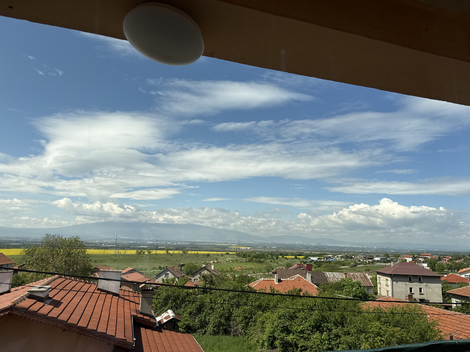 The image shows an overview of a suburban area with red-tiled roofs in the city of Sofia, Bulgaria, viewed through a window. In the background, there is a mountain range, likely the Vitosha Mountain. In the middle ground, there are yellow fields. Power lines are also visible, as well as structures indicating city infrastructure. The sky is blue with scattered clouds. A light fixture and part of the window frame are visible at the top of the frame.