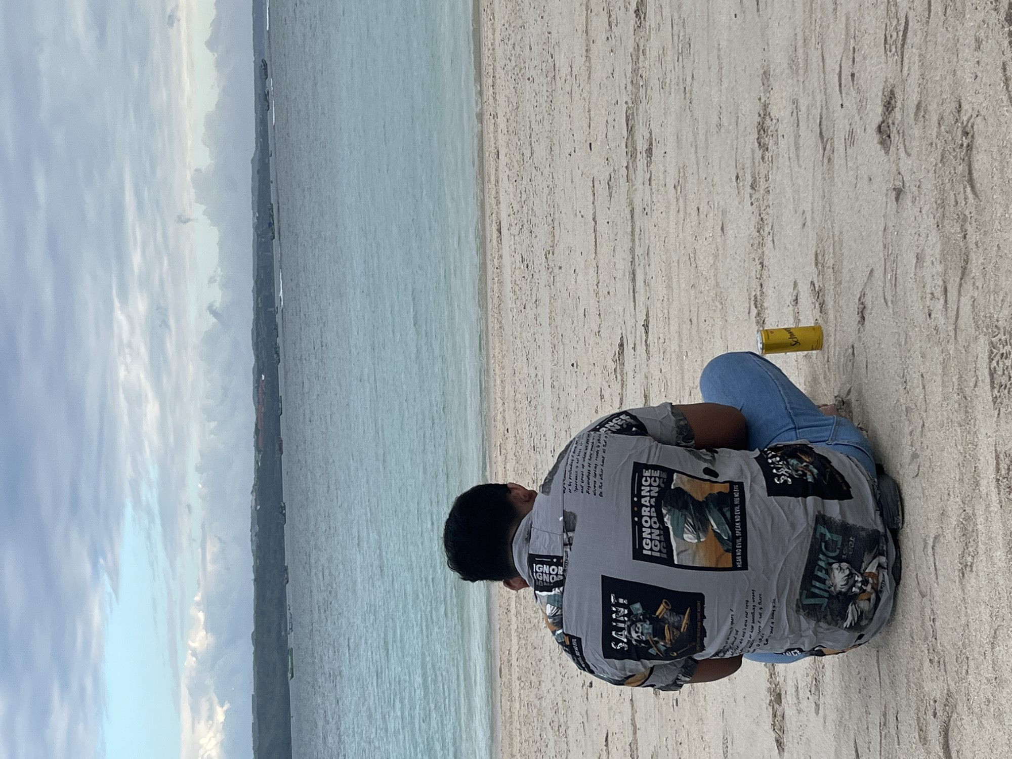 A person is sitting on a sandy beach in Kuta, Indonesia, facing a calm, turquoise ocean. The person is wearing a grey shirt with a graphic print and blue jeans, and is seated with their legs crossed. A yellow can of what appears to be Schweppes is placed on the sand to their right. The sky is overcast with scattered clouds, and a distant coastline with vegetation is visible across the water. Several boats are scattered on the ocean surface. The overall scene depicts a relaxed moment by the sea.