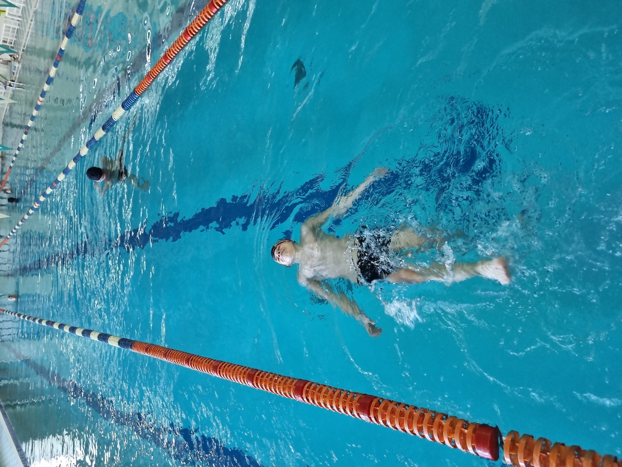 A high-angle shot of a swimming pool shows two individuals swimming. The pool is divided into lanes by floating lane dividers, alternating orange and white/blue sections. One swimmer is farther away, partially submerged with only their head visible. The closer swimmer is executing a backstroke. They are wearing a black swimsuit and a cap. Ripples and splashes indicate movement in the water. The pool water appears turquoise. The location is an indoor swimming pool of undetermined city.