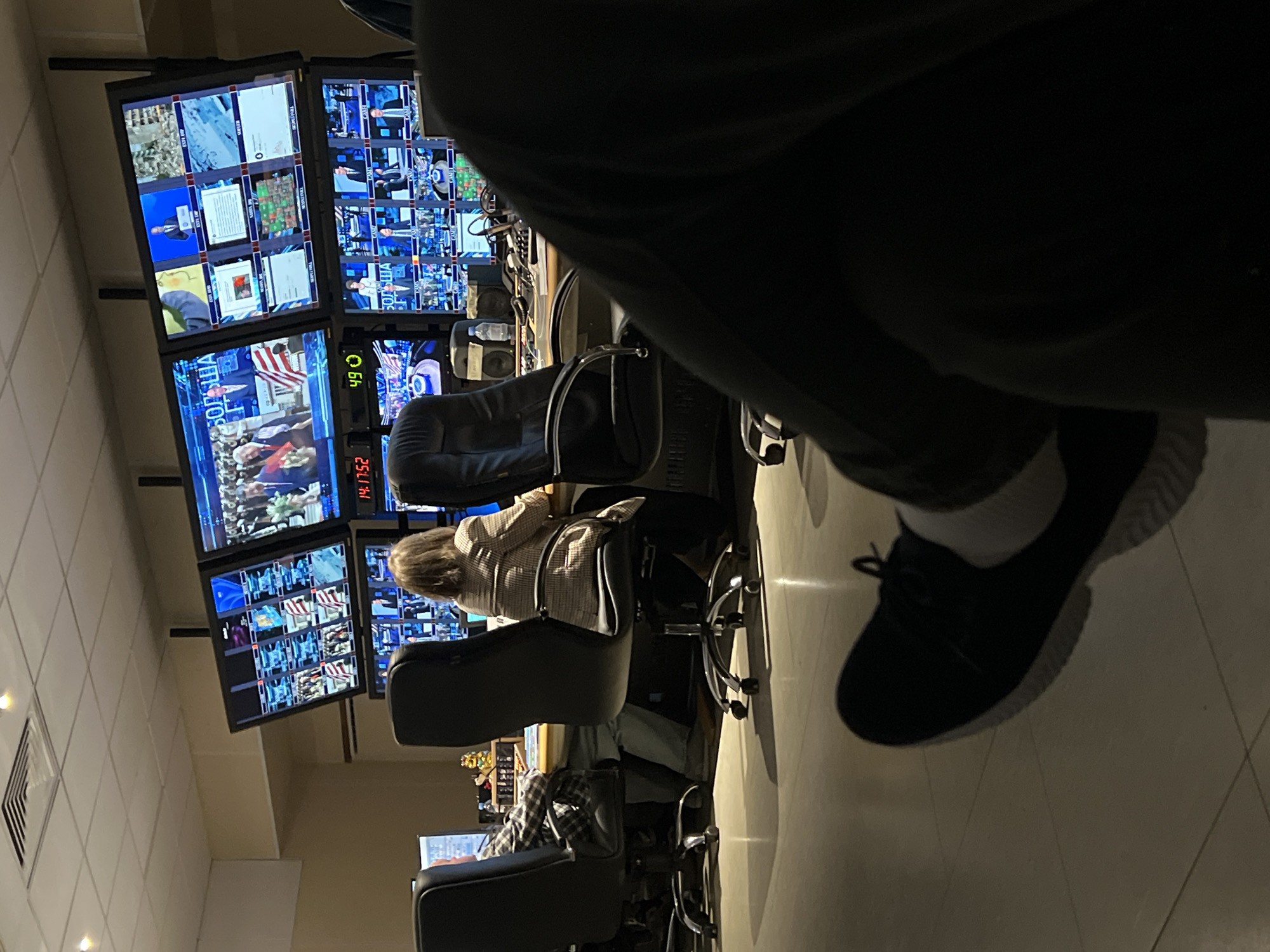 The image depicts the interior of a video control room or newsroom, possibly a production studio, with several monitors displaying various feeds. Multiple people are seated in office chairs, seemingly working in front of computers or control panels. A person's leg and foot, wearing a dark shoe with a light-colored sole and a white sock, are prominently featured in the foreground. The location and city (Khimki, Russia) cannot be verified from the visual data provided.