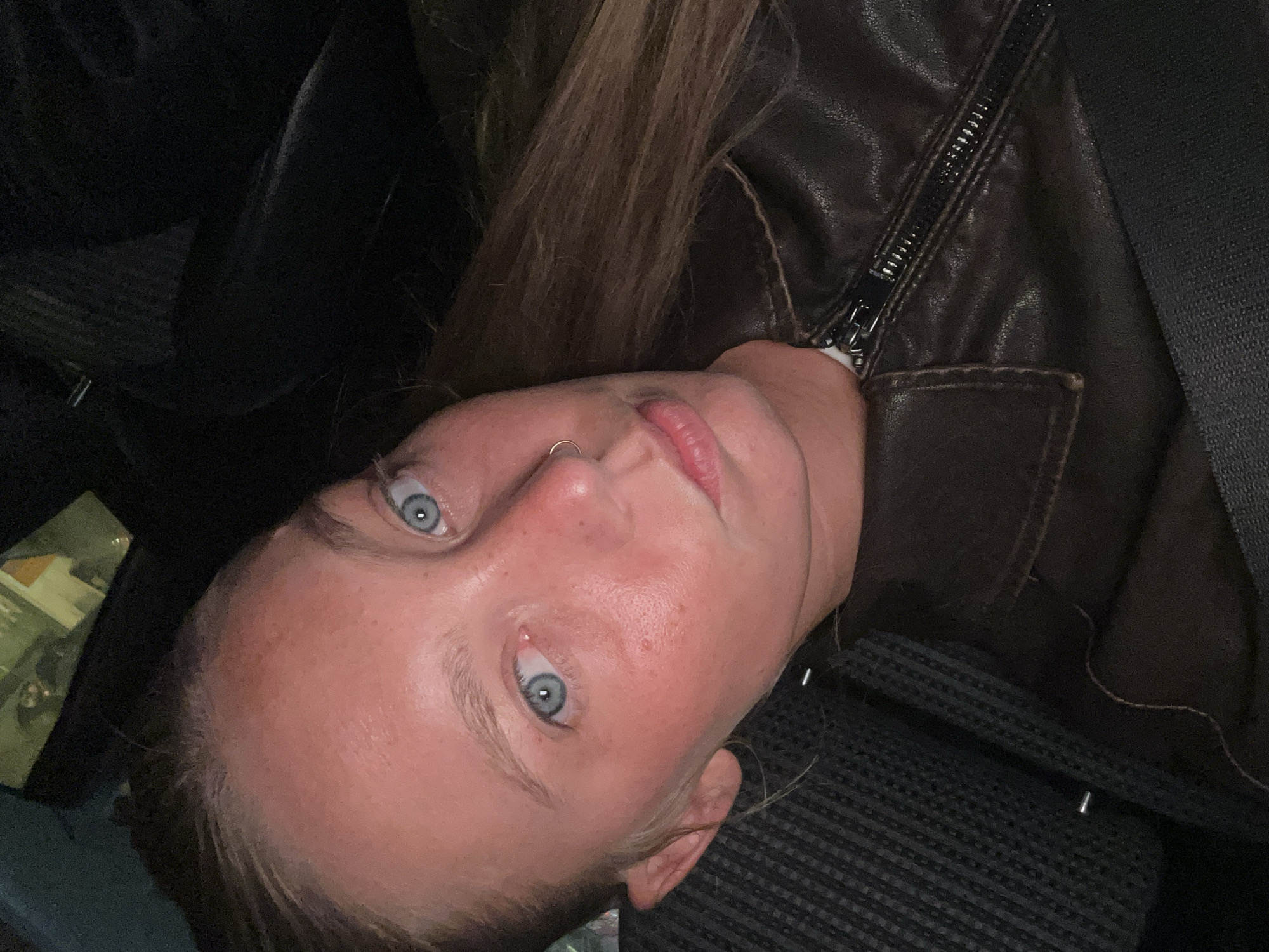 A selfie of a fair-skinned woman with blue eyes and brown hair. She is wearing a brown leather jacket and a seatbelt. She appears to be inside a vehicle. There is no indication in the image that places her in Noisy-le-Grand, France.
