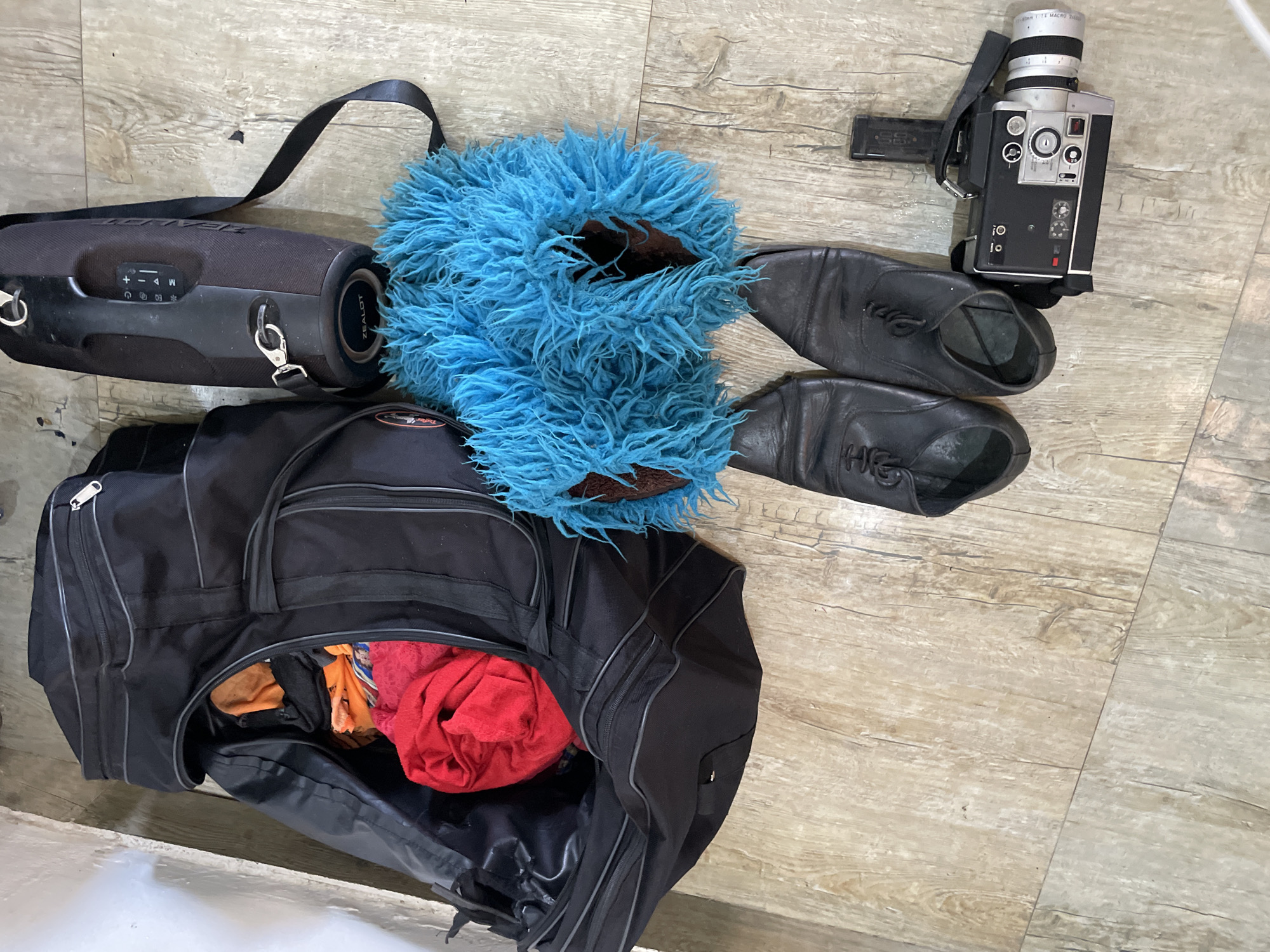 The image depicts a collection of items arranged on a light-colored, wood-patterned floor, likely indoors. The scene is located in Ado, Nigeria.
Central to the composition is a large, open black duffel bag, partially filled with brightly colored fabrics, including a prominent red item. To the right of the duffel bag is a large, black cylindrical speaker with a handle and straps. Below the speaker and partially obscuring it is a fluffy, bright blue accessory, possibly part of a costume or prop.
In front of the blue accessory and slightly to the left are a pair of black leather dress shoes. To the right of the shoes and closer to the bottom right of the frame is a vintage-style video camera, silver and black in color, with a prominent lens and strap. The lighting suggests an indoor environment, and the overall arrangement of objects implies they may have been recently used or are being prepared for transport or storage. There are no people visible in the image.