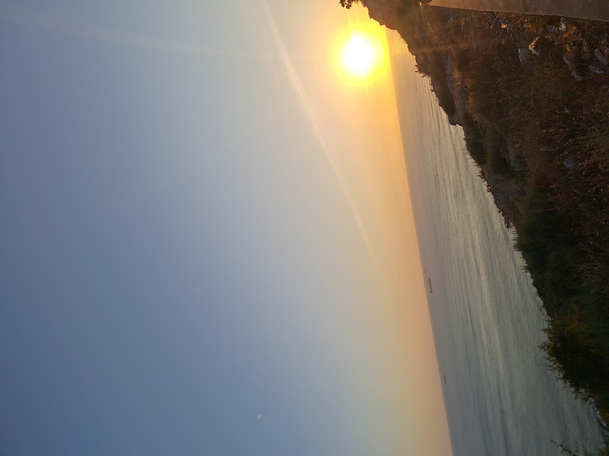 The image, taken in Safi, Morocco, depicts a serene coastal landscape at either sunrise or sunset. The scene is viewed from a high vantage point, overlooking the ocean.
The dominant feature is the intensely bright sun, positioned in the upper right quadrant of the correctly oriented frame. It radiates a brilliant yellow-orange light that creates a dramatic gradient in the sky, transitioning from vivid orange around the sun to a soft, clear blue further to the left. A faint, small, bright disc is noticeable in the lower-middle left section of the blue sky.
The calm expanse of the ocean occupies the central and right-central areas of the image. The water reflects the warm light of the sun, creating a glimmering path on its surface, and shows subtle ripples. Several dark, indistinct outlines, appearing to be distant ships or boats, are visible on the water, particularly in the central and lower right portions.
Along the far right edge, a rugged, dark coastline or cliff descends towards the sea. This landmass is covered with sparse, dry vegetation and visible rocky patches. Some scattered debris or litter can be observed on the upper part of the hillside, near the top right corner.
No people or specific activities are visible in the photograph, highlighting the natural beauty and tranquility of the coastal environment during this illuminated time of day. There is no discernible text present in the image.