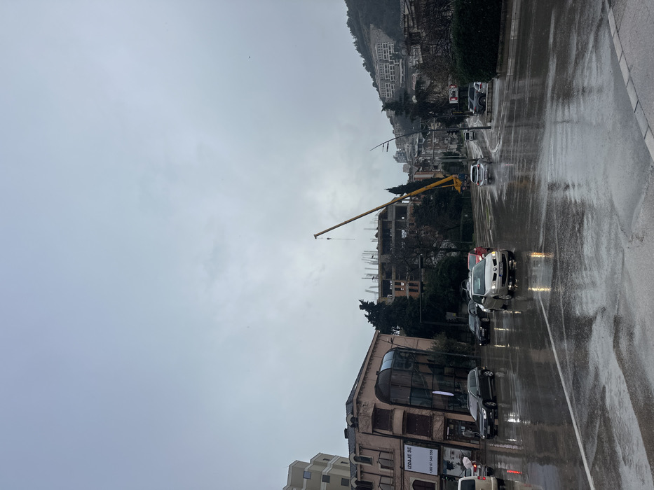The image depicts an urban street scene in Budva, Montenegro, specifically located on Mediteranska Street, near its intersection with Prve Proleterske. The environment is characterized by an overcast, light gray sky and wet asphalt roads, indicating recent rain or drizzle, with reflections visible on the road surface.
In the foreground, a multi-lane road features active vehicle traffic. A silver sedan occupies the center-foreground, moving away from the viewer. To its left, a dark-colored SUV is also heading away, and a white van is visible on the far left. A pedestrian holding a white umbrella is partially visible near the white van.
On the left side of the frame, a multi-story building with a light pink/peach facade features a distinctive curved glass corner. A commercial sign "IZDAJE SE" (FOR RENT) is visible on its side, along with the contact number "+382 67 549 556". Behind this structure, a multi-story concrete building is under construction, displaying exposed rebar. Adjacent to this construction site stands a large yellow mobile crane with its boom extended upwards, a hook dangling.
The right side of the frame reveals a steep hillside densely populated with multi-story residential buildings. Various trees and shrubbery are interspersed throughout the scene.