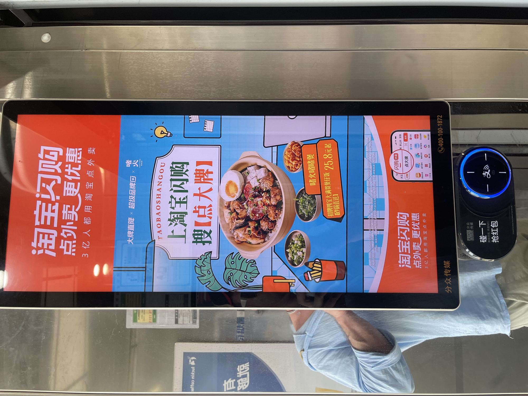 The image shows a digital advertising screen in Panyu District, China. The screen displays an advertisement for "Taobao Dian Gou" (淘宝闪购), which is a food delivery service. The advertisement features images of food and text in Chinese, promoting discounts and special offers. Below the main screen, there is a touch-sensitive button with a blue light ring, likely for user interaction. The surrounding environment appears to be an outdoor or semi-outdoor public space, possibly near a transportation hub or commercial area, given the metallic fixtures and the presence of other advertisements. The overall scene is commercial and technological, focused on promoting online shopping and food delivery services.