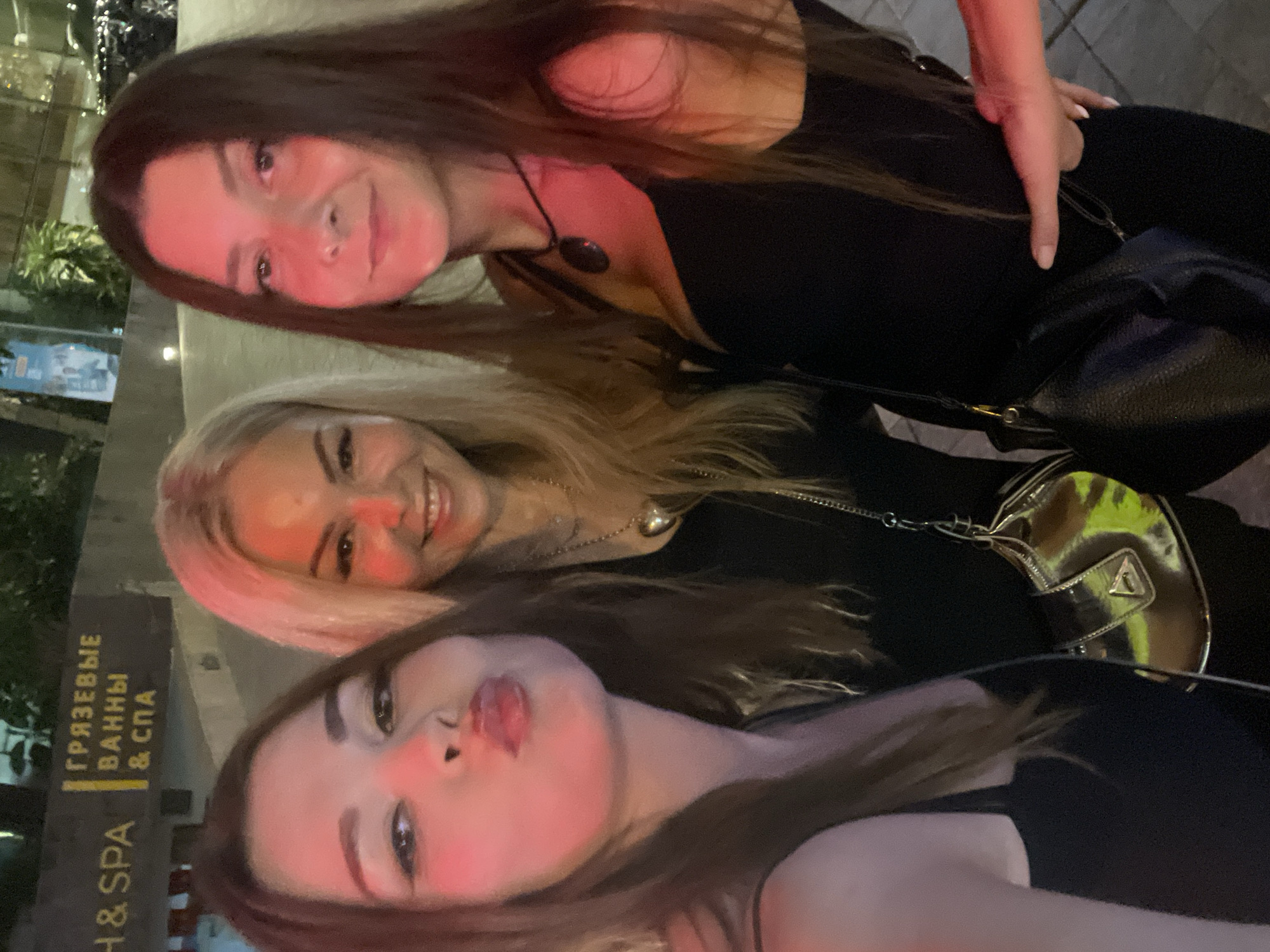 The image shows three young women posing for a selfie. They are all smiling and appear to be in good spirits. The woman in the foreground is sticking her tongue out. The woman in the middle has blonde hair and is wearing a black top. The woman in the back has dark hair and is also wearing a black top. They are outdoors, and it appears to be nighttime, as evidenced by the artificial lighting. The scene is likely in a public place, possibly a street or an outdoor seating area of a restaurant or bar, given the presence of a sign in the background. The sign has text in Cyrillic and English. The Cyrillic text reads "ГРЯЗЕВЫЕ ВАННЫ & СПА" which translates to "Mud Baths & Spa". The English text reads "H & SPA". The overall mood is casual and fun, suggesting a night out with friends. The lighting is somewhat dim, with a pinkish hue in some areas, possibly from ambient lighting or a flash.