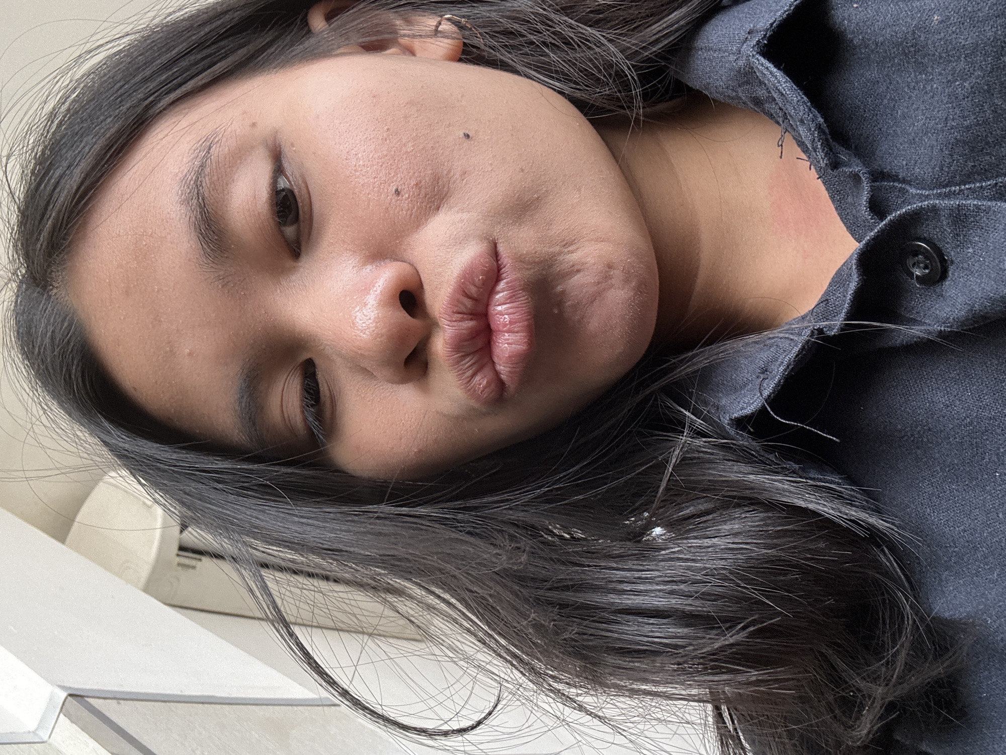 A close-up, selfie-style, eye-level shot captures a young Indonesian woman with dark hair and medium-toned skin puckering her lips. Her hair is dark and appears slightly windswept, framing her face. She is wearing a dark collared shirt. The background is out of focus but appears to be an outdoor urban environment, possibly a walkway or building exterior in East Jakarta, Indonesia, suggested by the style of the architecture and ambient lighting. The image is clear and well-lit, focusing on the woman's facial expression and upper body.