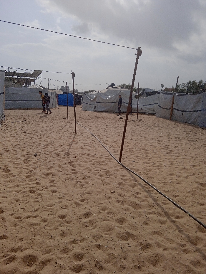 The image depicts an outdoor encampment on sandy terrain in Khan Yunis, Palestinian Territory, under a cloudy sky. The ground is light brown sand, densely covered with footprints. Multiple makeshift shelters, primarily constructed from white tarpaulin or fabric, are arranged across the scene. Some tents are rounded or dome-shaped, while others are linear. A blue tarp structure stands in the middle-ground. In the background to the left, a dark, angled structure, potentially solar panels, is visible above a tent, and distant buildings are faintly discernible.
Three individuals are present. On the left side of the mid-ground, two individuals walk away from the viewer towards the left. One wears a light-colored shirt and dark pants, carrying a dark backpack. The other wears a darker shirt and light pants. On the right side of the mid-ground, a single individual stands facing left near a tent, dressed in dark clothing.
Several wooden poles are vertically embedded in the sand, supporting dark wires or cables strung horizontally across the upper portion of the scene. A thick, dark cable also lies on the sandy ground, extending from the foreground into the middle distance. Dark, circular objects, consistent with water tanks, are visible atop some background tents, and a satellite dish is faintly seen above a tent on the right.
