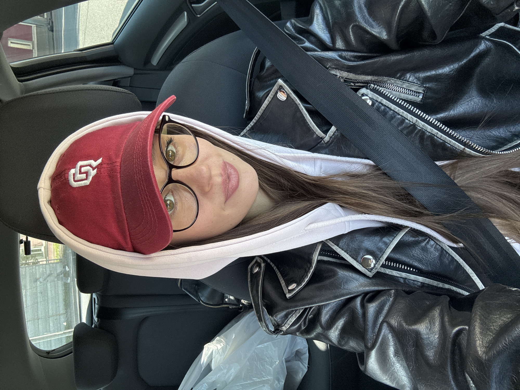 A woman is in the passenger seat of a car. She is wearing glasses, a red baseball cap with a white "OU" logo, and a white hooded sweatshirt under a black leather jacket. She is wearing a seatbelt. The interior of the car is visible. The location is unknown; there is no indication of Krasnoyarsk, Russia.