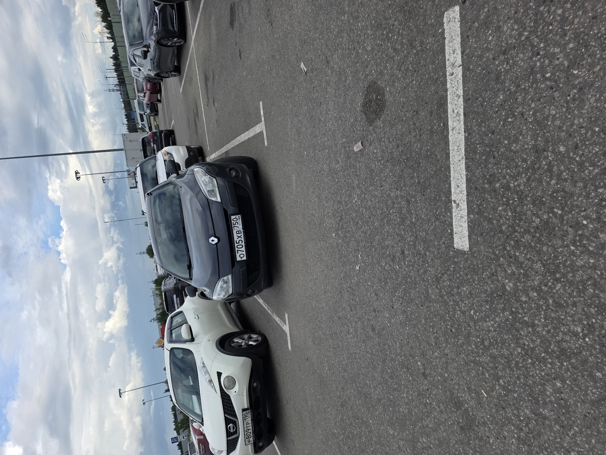 A black Renault Megane is parked in a lot. The car has a license plate that reads "0705XB750". To the left of the Renault, a white Nissan Juke is parked. The Nissan has a license plate that reads "P809TP190". The parking lot is paved with asphalt and marked with white lines. There are other cars parked in the background, and a cloudy sky is visible above.