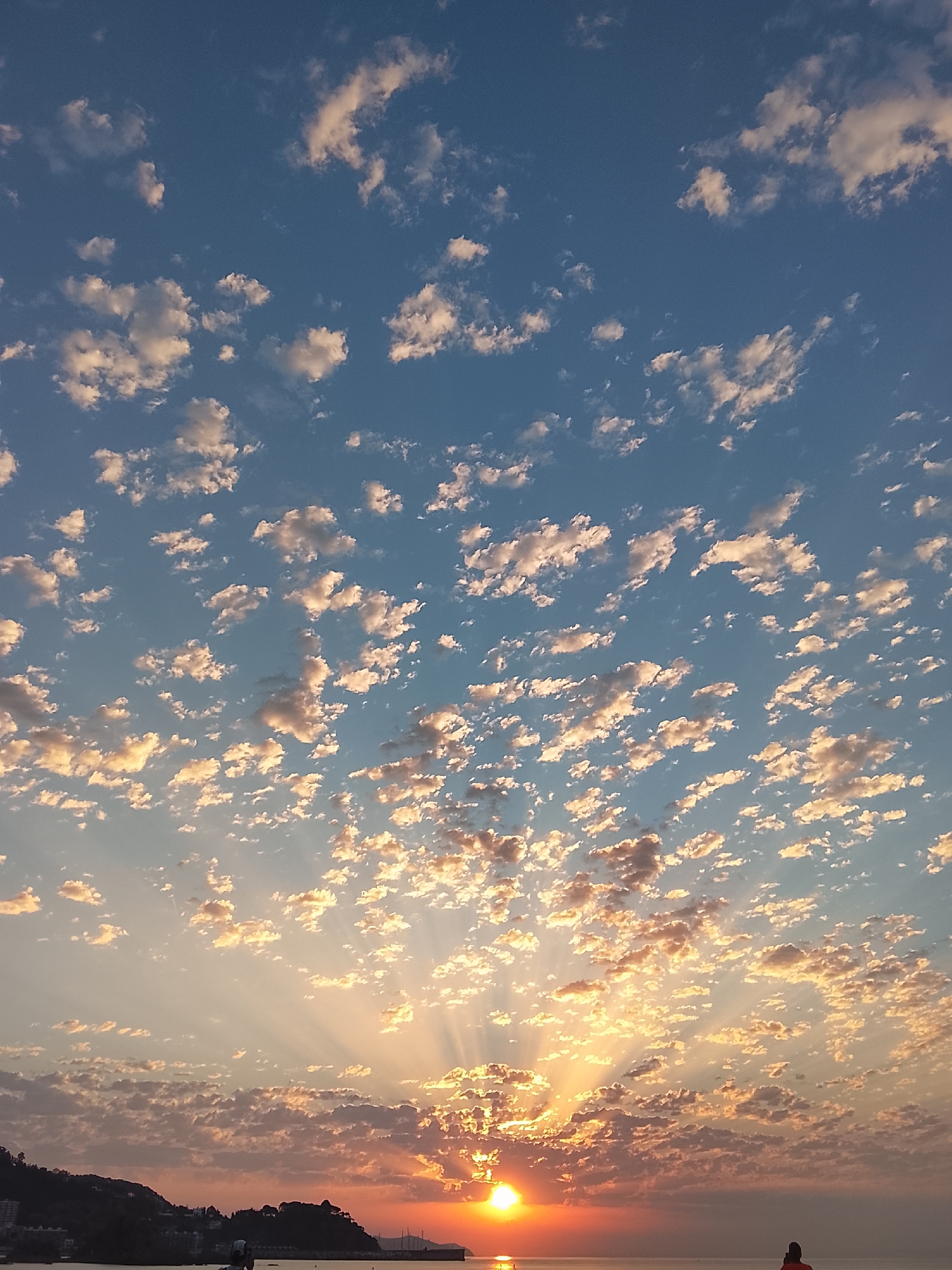 This is a wide-angle, full-frame photograph capturing a sunset over the sea in Blanes, Spain. The sky is filled with scattered cumulus clouds, tinged with warm hues of orange and yellow near the horizon, transitioning to a clear blue overhead. Rays of sunlight emanate from behind the clouds, creating a dramatic lighting effect. In the foreground, silhouetted against the sunset, is a dark, hilly coastline with buildings visible on the slopes. A body of water, likely the Mediterranean Sea, occupies the lower portion of the image. Two individuals are present in the scene. One is a dark silhouette of a person in the lower right corner, facing away from the camera. Another dark silhouette of a person is visible near the left edge of the image, also facing away. The overall mood is serene and picturesque, characteristic of a coastal sunset.