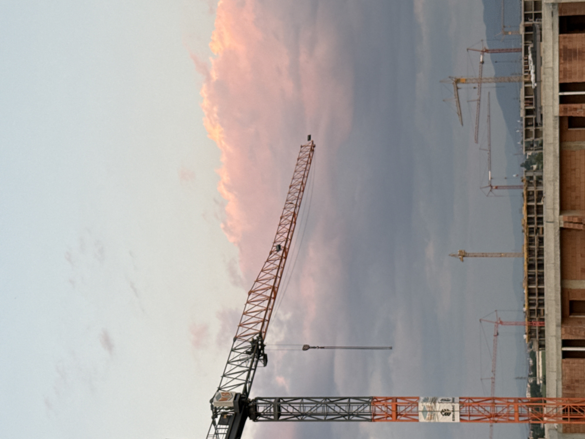 The image shows a construction site in Plovdiv, Bulgaria. Multiple cranes are visible against a sky with some pink clouds. A building under construction is on the right side of the image.